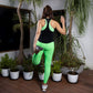 A woman in a solid black top with racer back stretches her leg on a wooden deck, surrounded by potted plants.