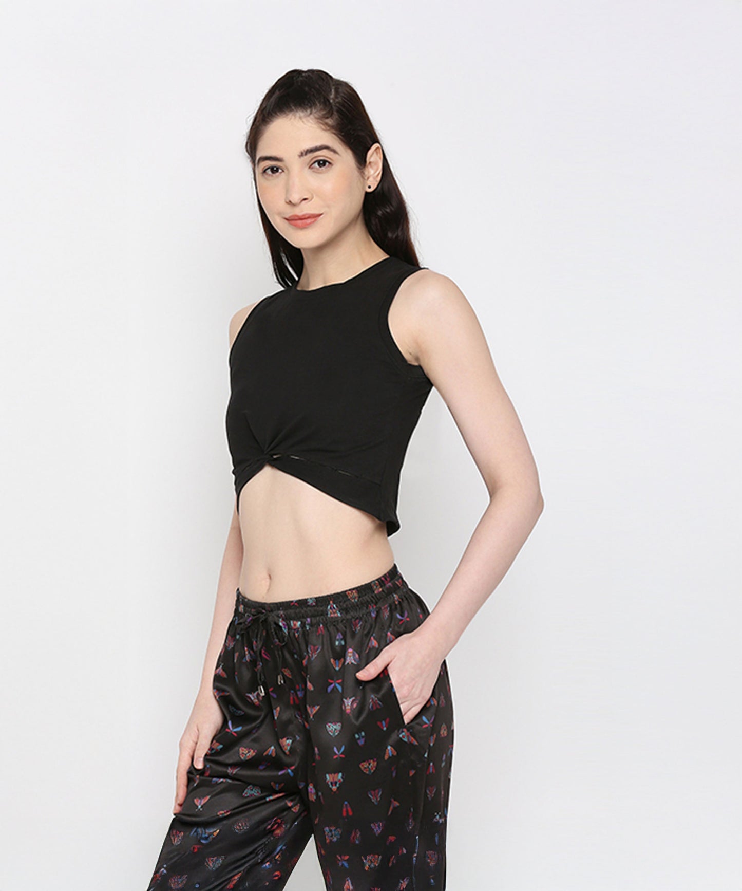 Woman wearing the Black Twisted Crop Top—a black sleeveless, round neck crop top with a twist pattern—paired with printed black pants, stands stylishly with one hand in her pocket against a white background.