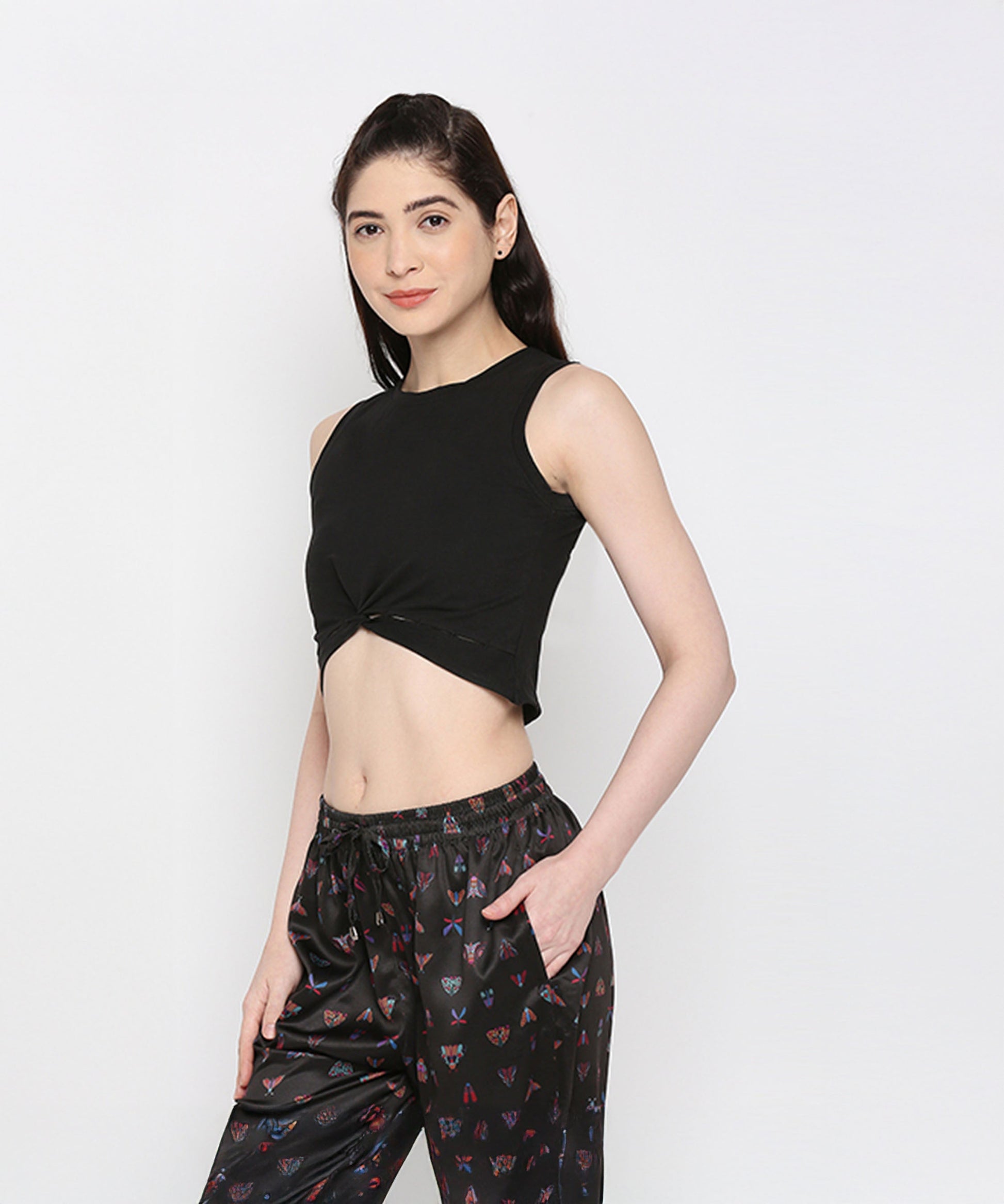 Woman wearing the Black Twisted Crop Top—a black sleeveless, round neck crop top with a twist pattern—paired with printed black pants, stands stylishly with one hand in her pocket against a white background.