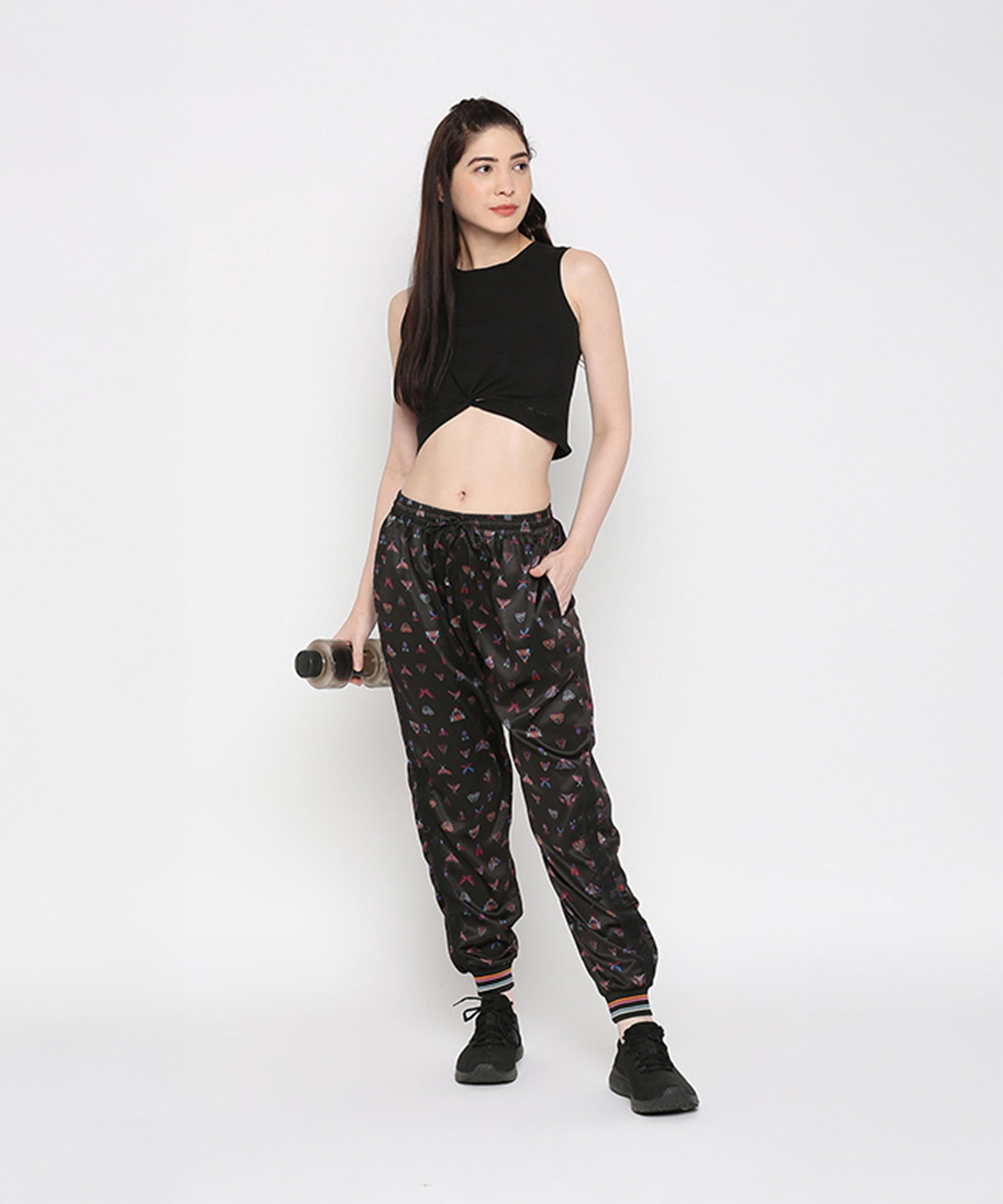 A woman poses against a white background wearing a Black Twisted Crop Top, printed joggers, and sneakers while holding a water bottle.