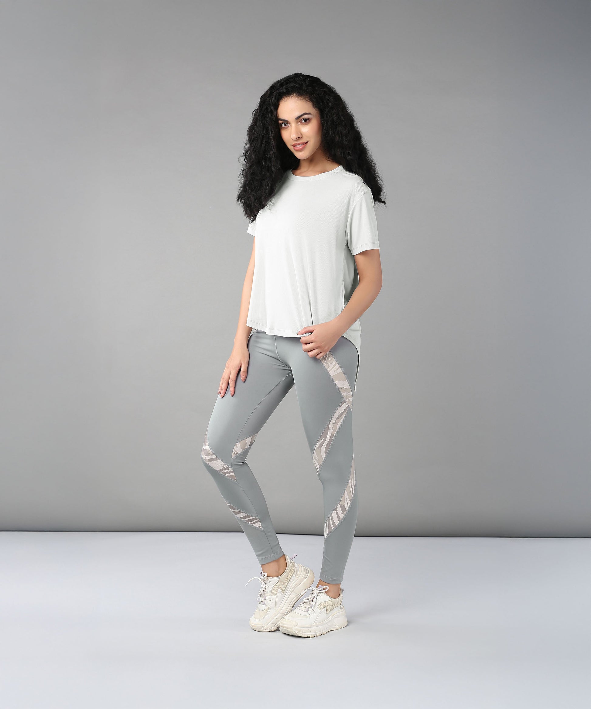 Woman with long curly hair wears the Grey Top with Patched Legging, made from super soft, moisture-wicking fabric, paired with sneakers and poses against a plain gray background.