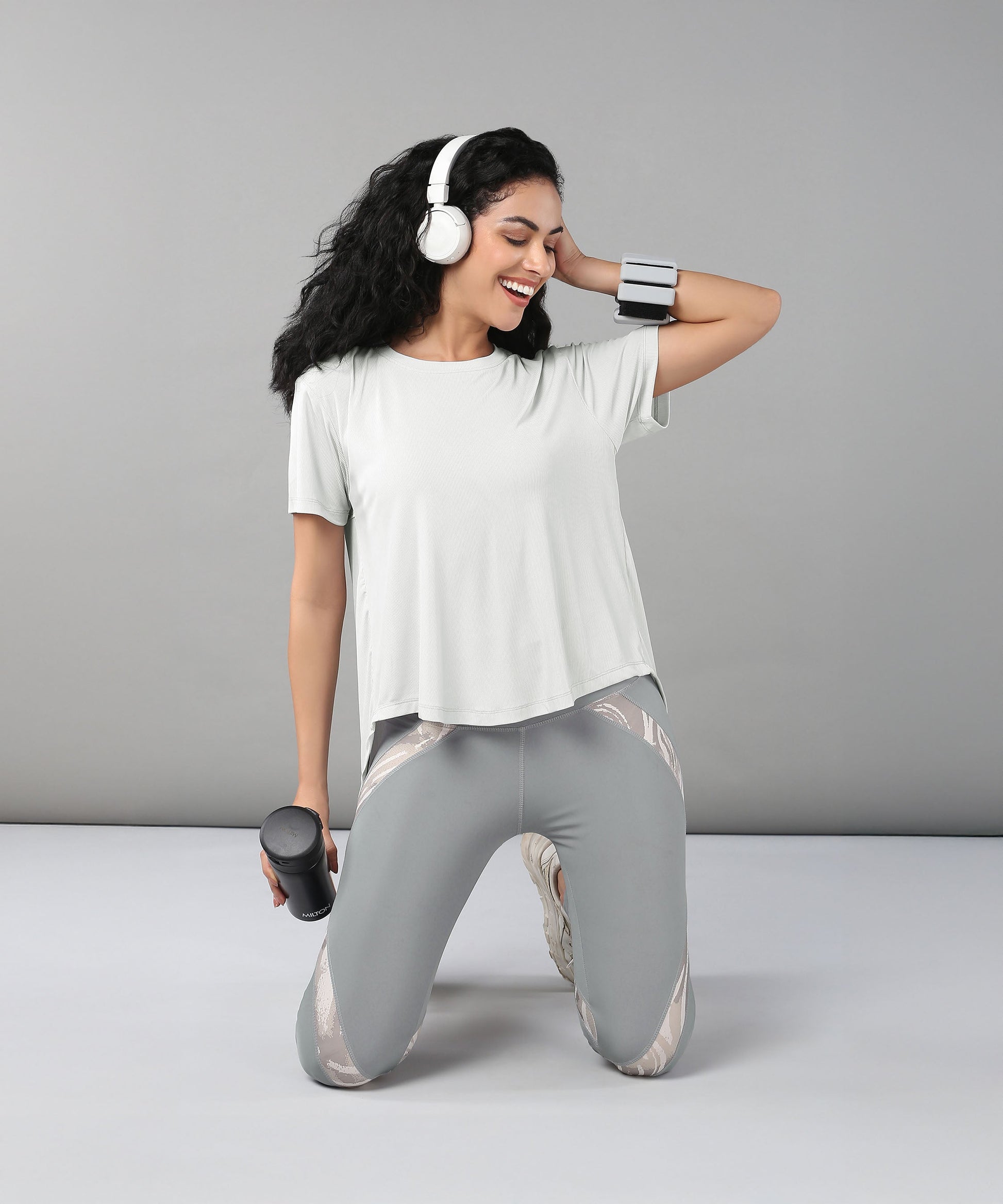Smiling woman kneeling with headphones and holding a water bottle, dressed in the Grey Top with Patched Legging—workout clothes made from super soft fabric for all-day comfort.