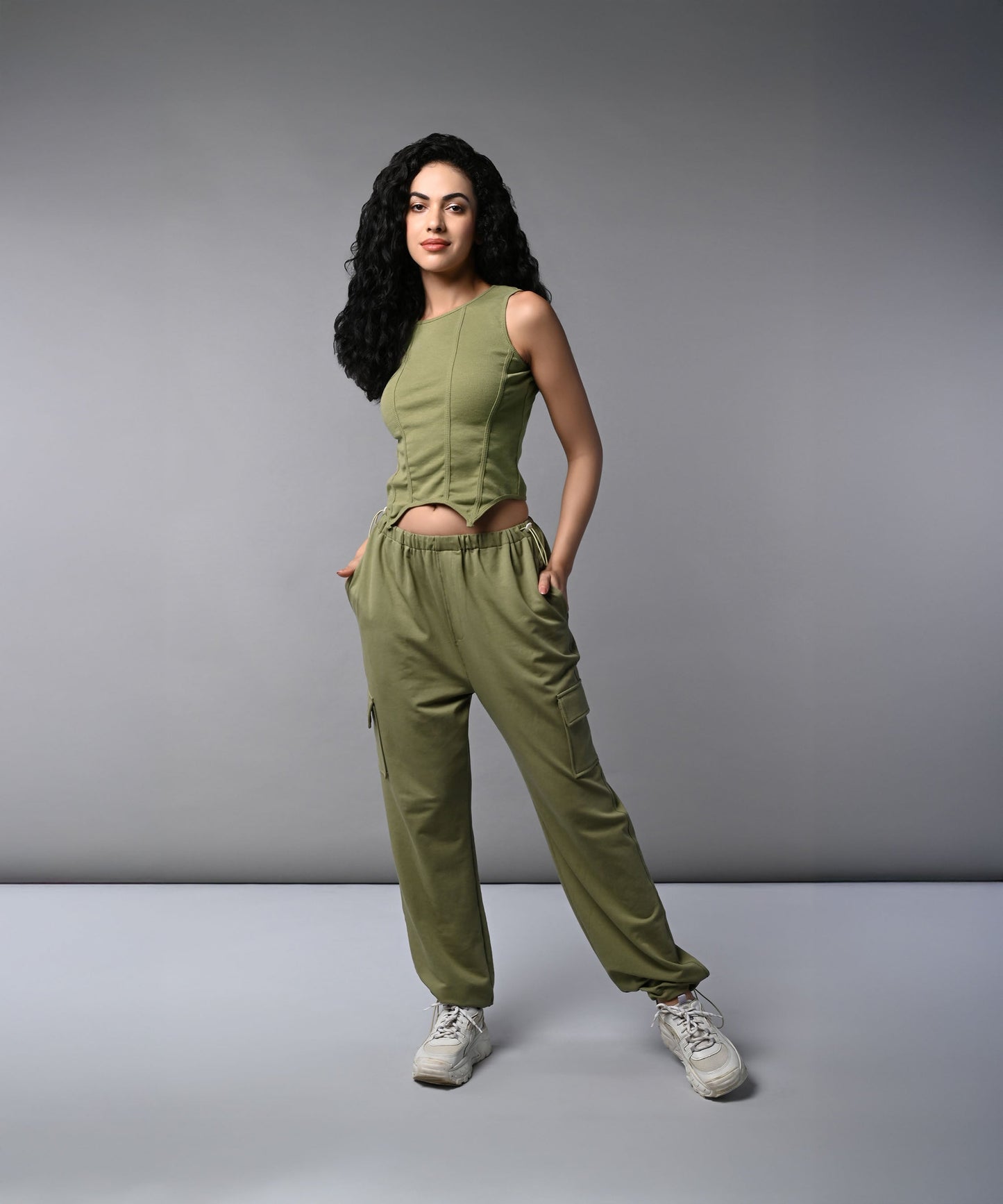 A woman with long dark hair models the Olive Corset Crop - Top with Parachute Pants, standing hands-in-pockets against a gray background.