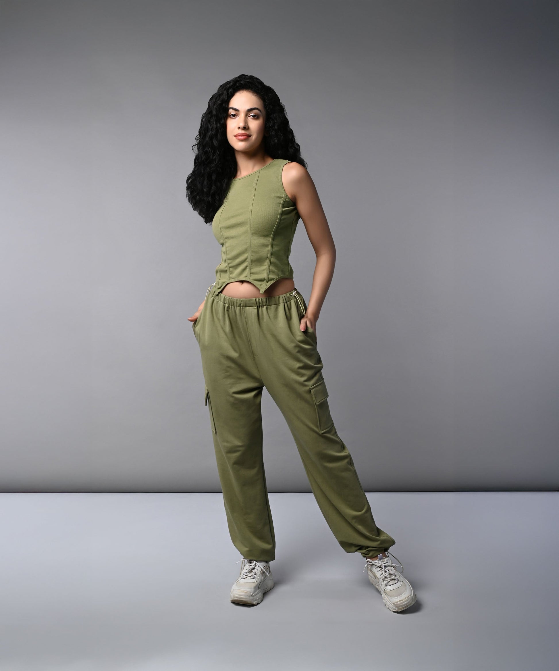 A woman with long dark hair models the Olive Corset Crop - Top with Parachute Pants, standing hands-in-pockets against a gray background.