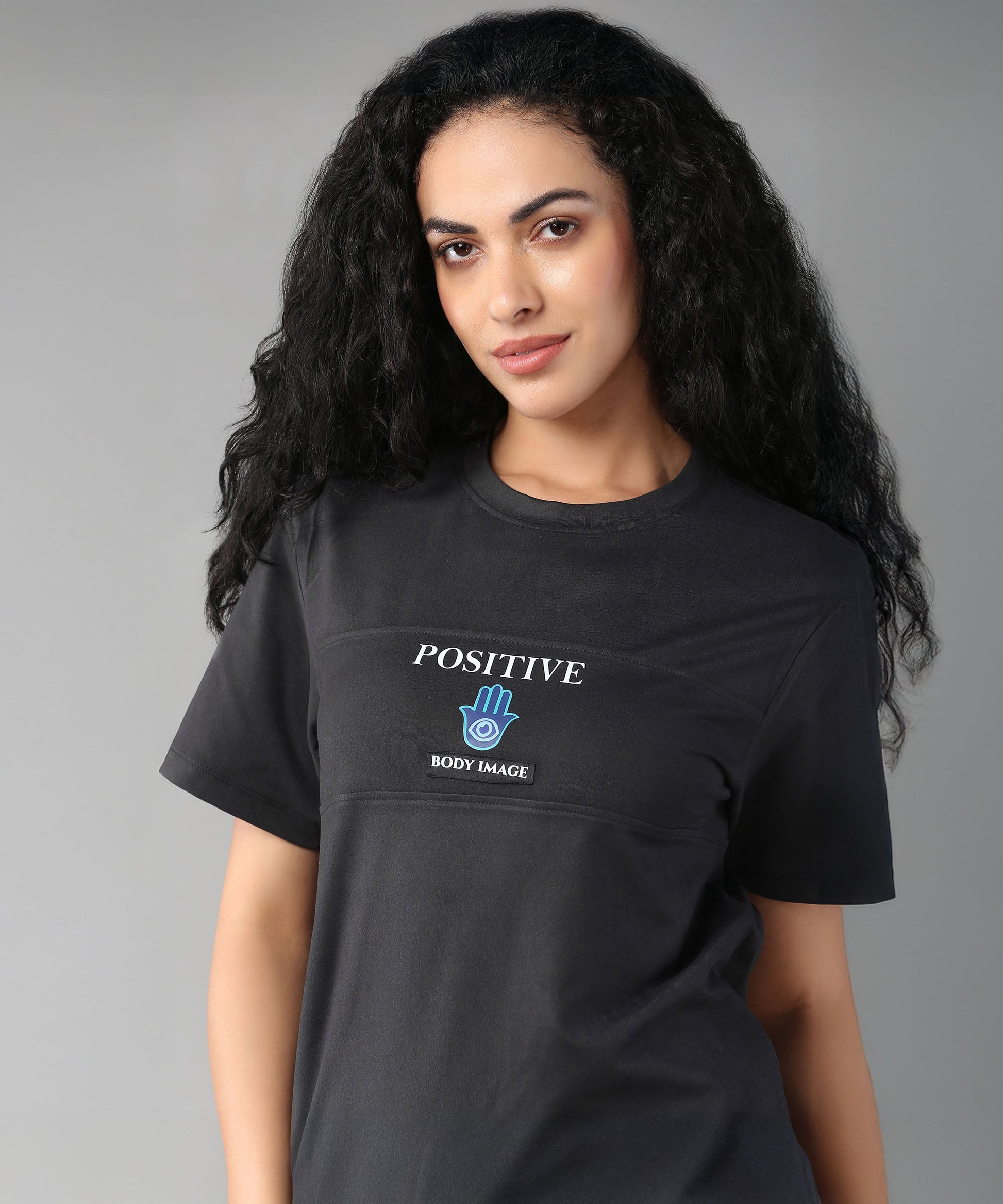 A woman with long curly hair smiles against a gray background, wearing the Positive Body Image T Shirt—a black, basic half-sleeve tee with a "Positive Body Image" print.