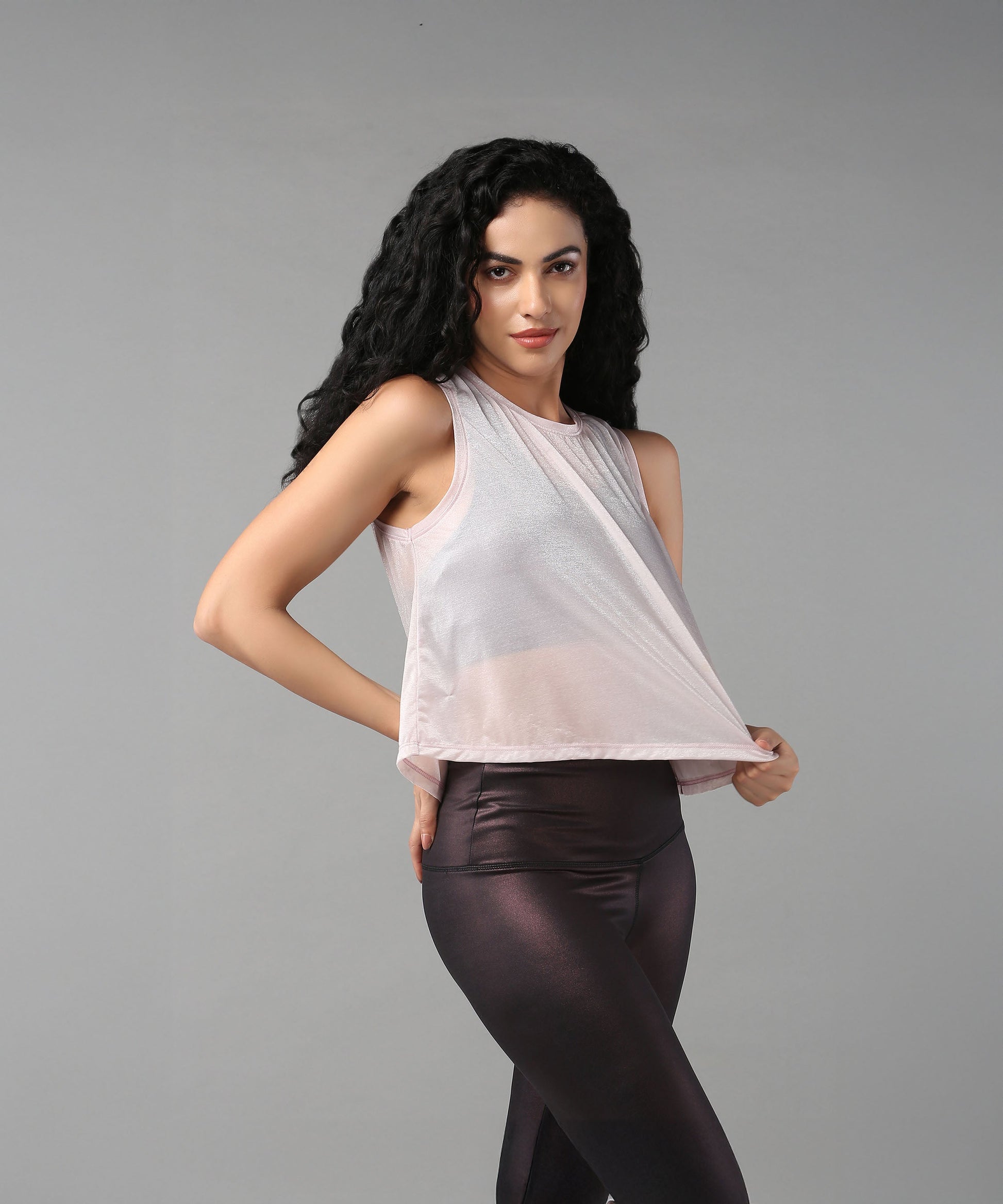 A woman with long curly hair poses against a gray background wearing the Powder Pink Sheer Top and shiny leggings.