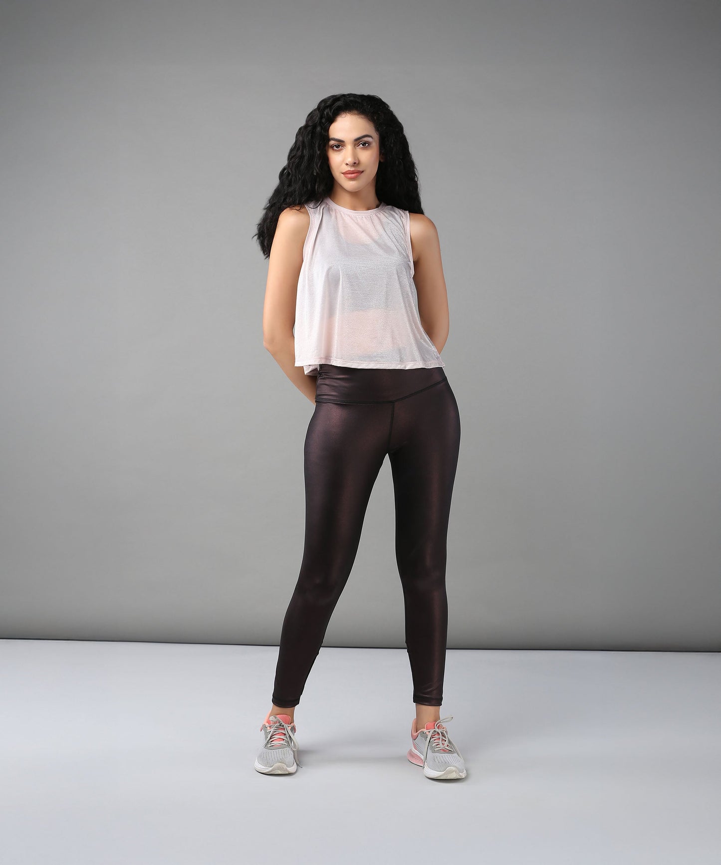 A woman with curly hair models the Powder Pink Sheer Top and shiny black leggings against a plain gray background.