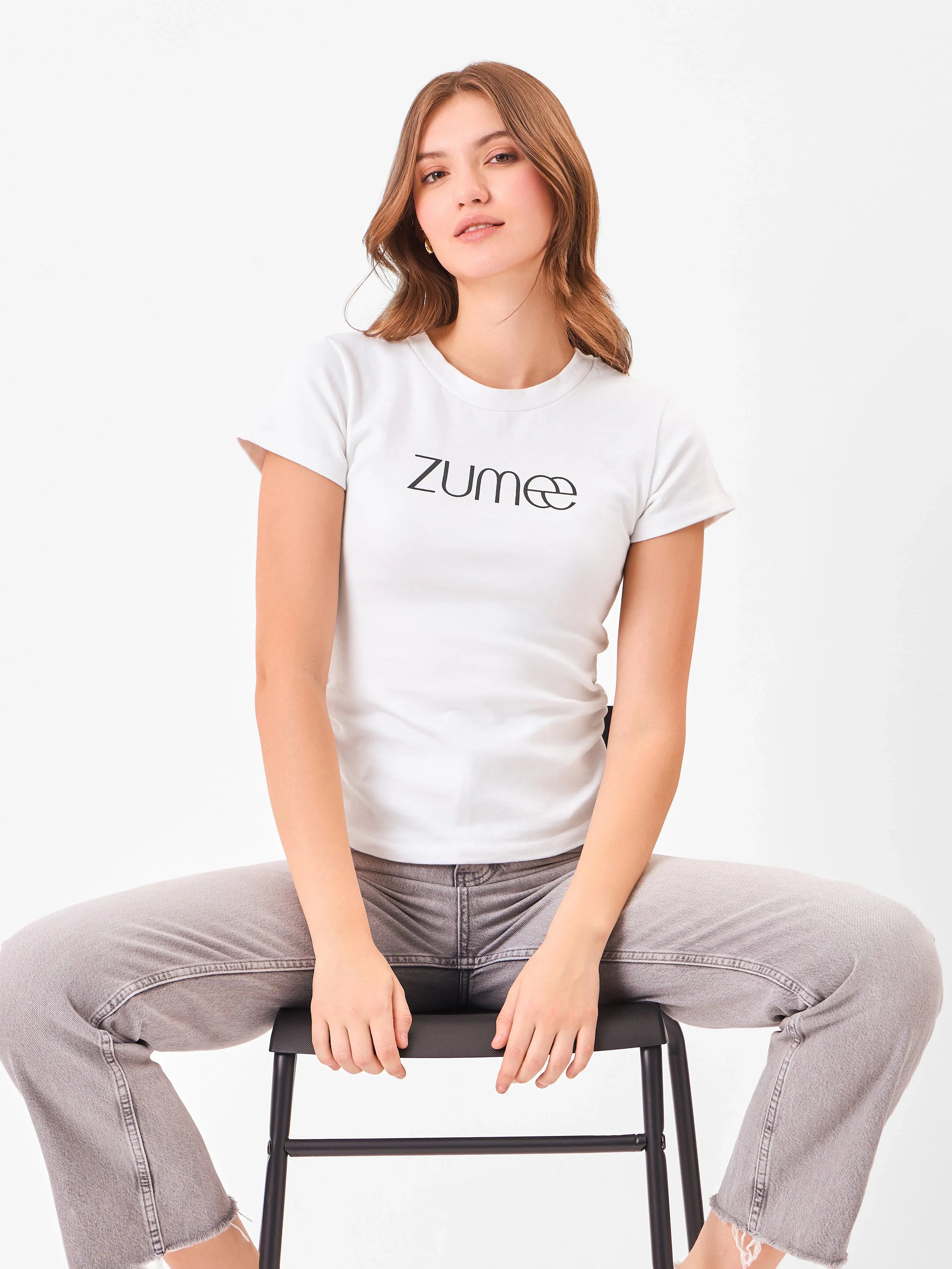 Female model wearing a white t-shirt with 'Zume' logo from Stylera, styling on a chair