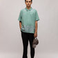 Stylera model wearing mint green casual men's fashion polo with subtle leaf print, black pants holding gray cap