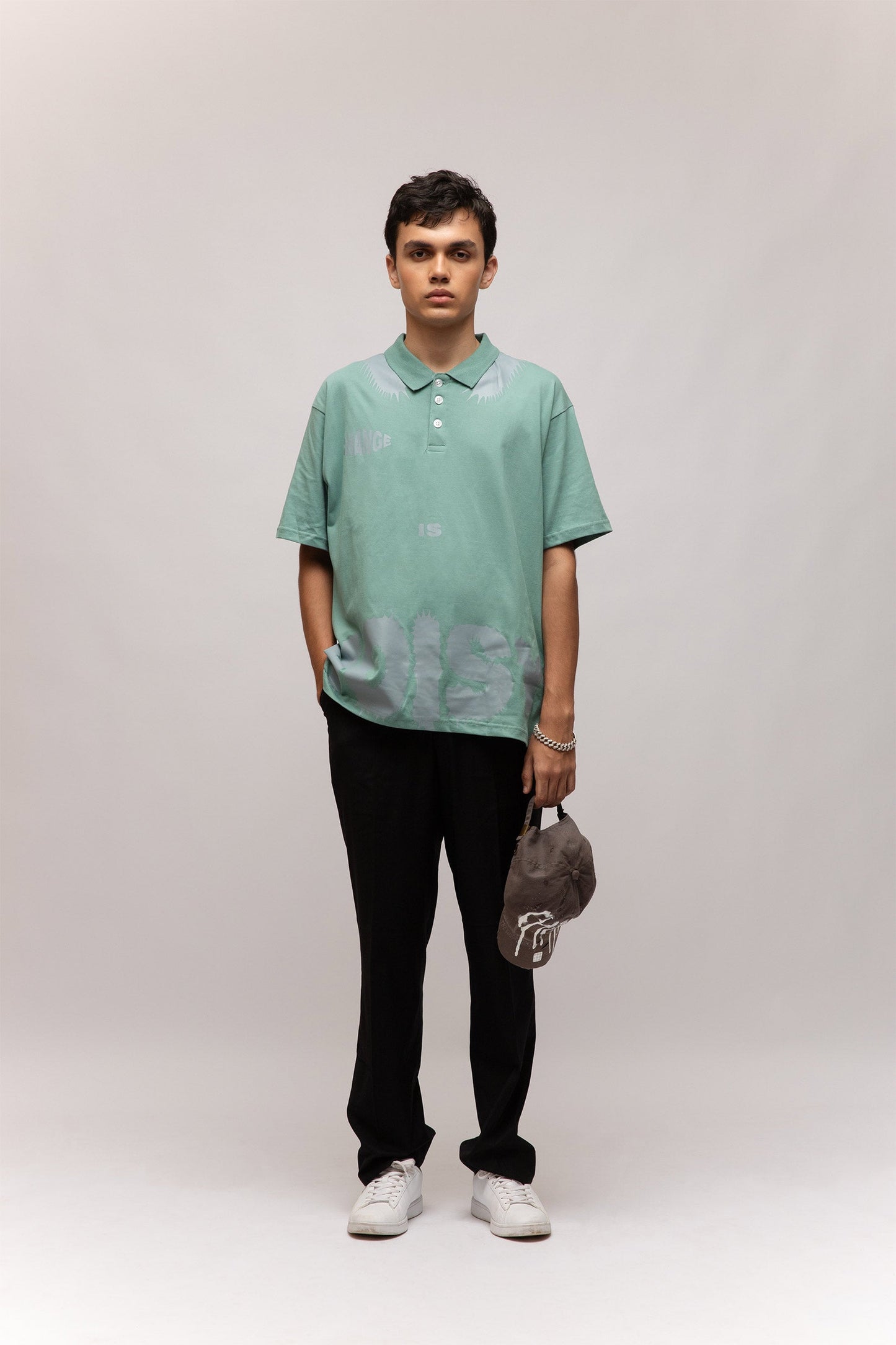 Stylera model wearing mint green casual men's fashion polo with subtle leaf print, black pants holding gray cap