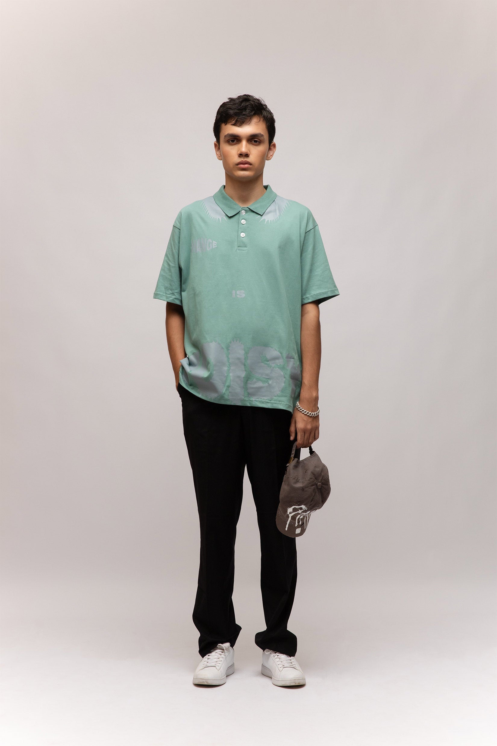 Stylera model wearing mint green casual men's fashion polo with subtle leaf print, black pants holding gray cap
