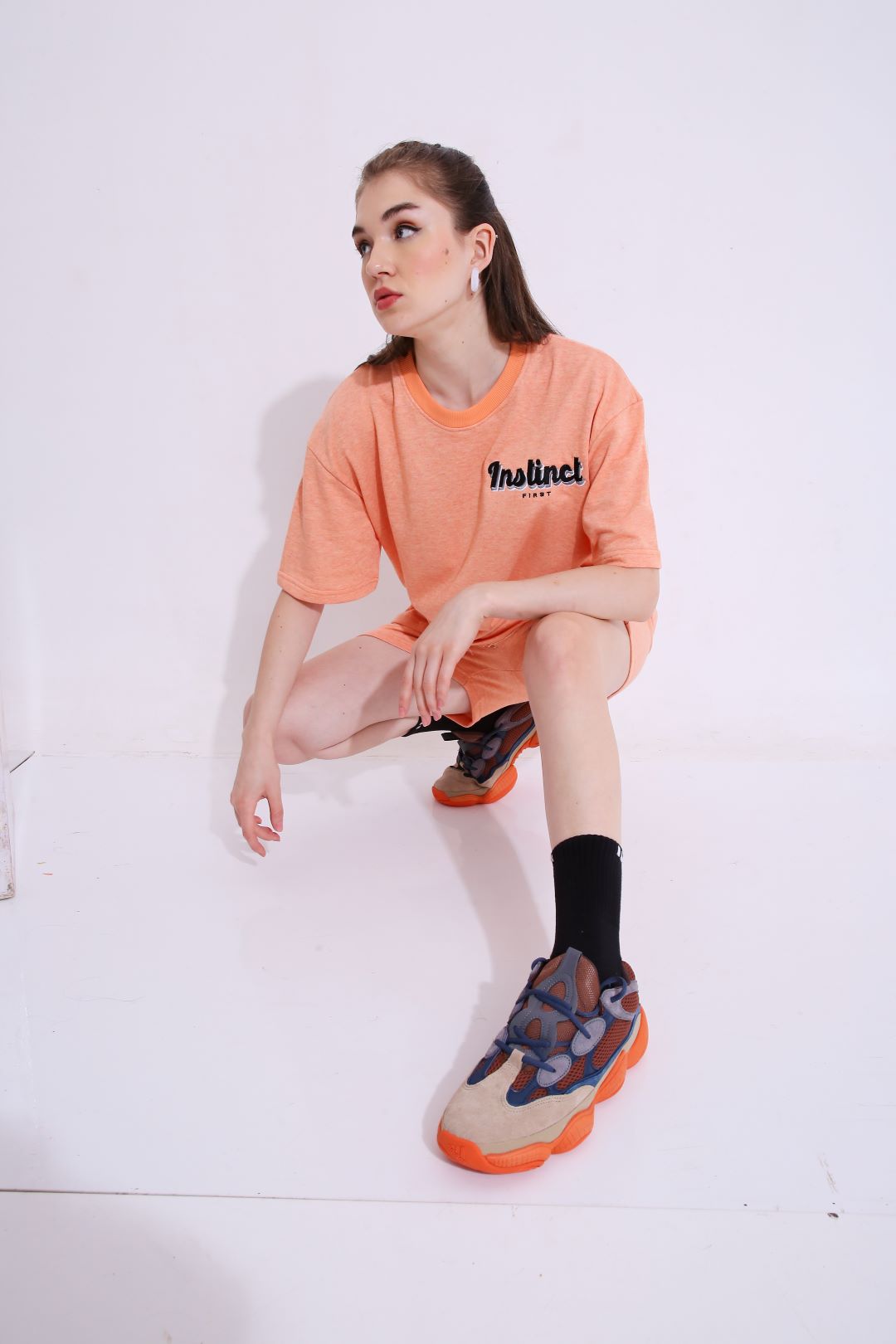 Young woman crouching in Stylera peach oversized tee with sneakers and black socks trendy oversized casual t-shirt