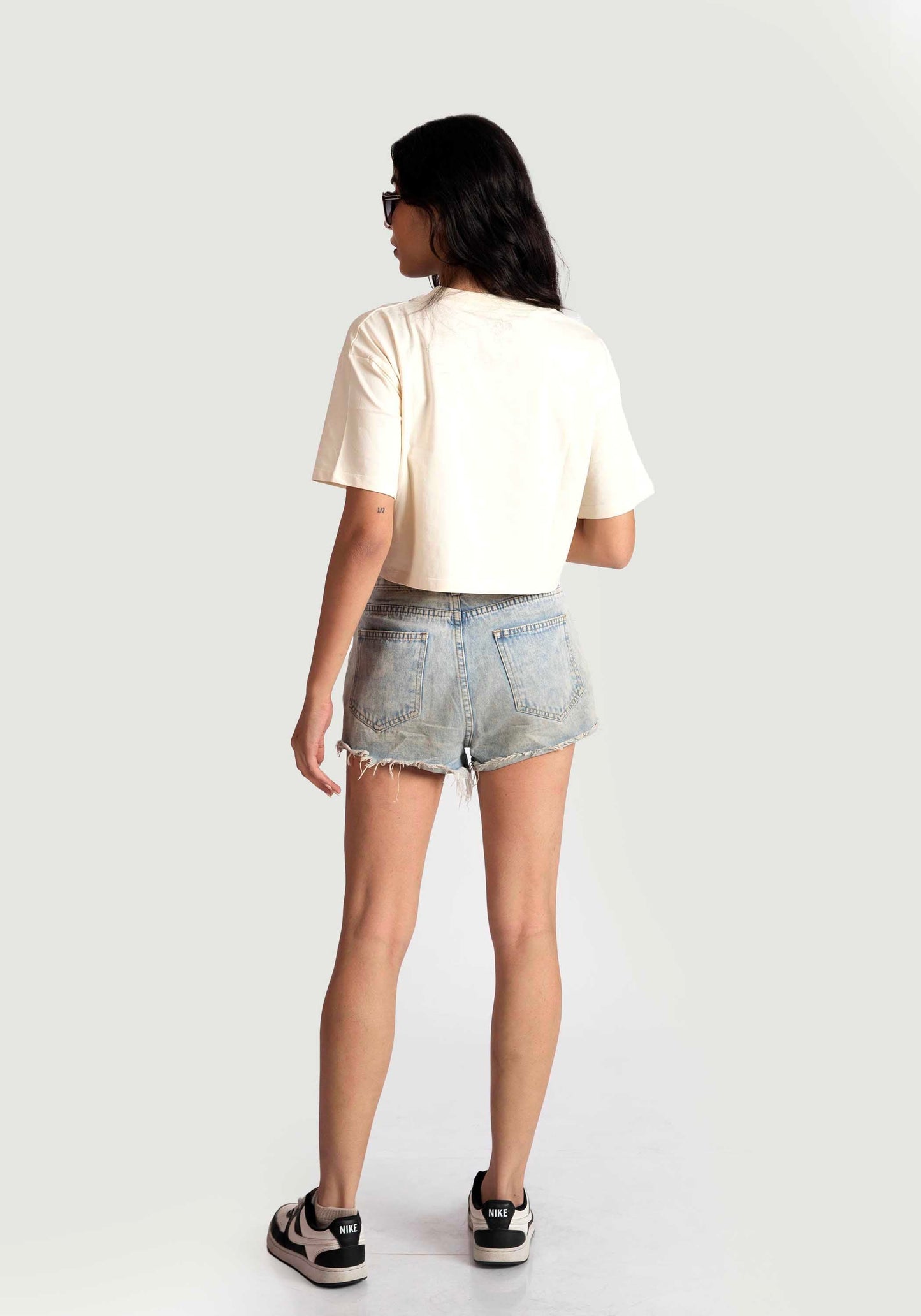 Woman wearing Stylera white oversized crop fit tee with light denim shorts and sneakers standing facing away
