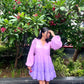 Model in women's purple gradient dress with puff sleeves and tiered skirt, white ankle boots, standing by tropical foliage