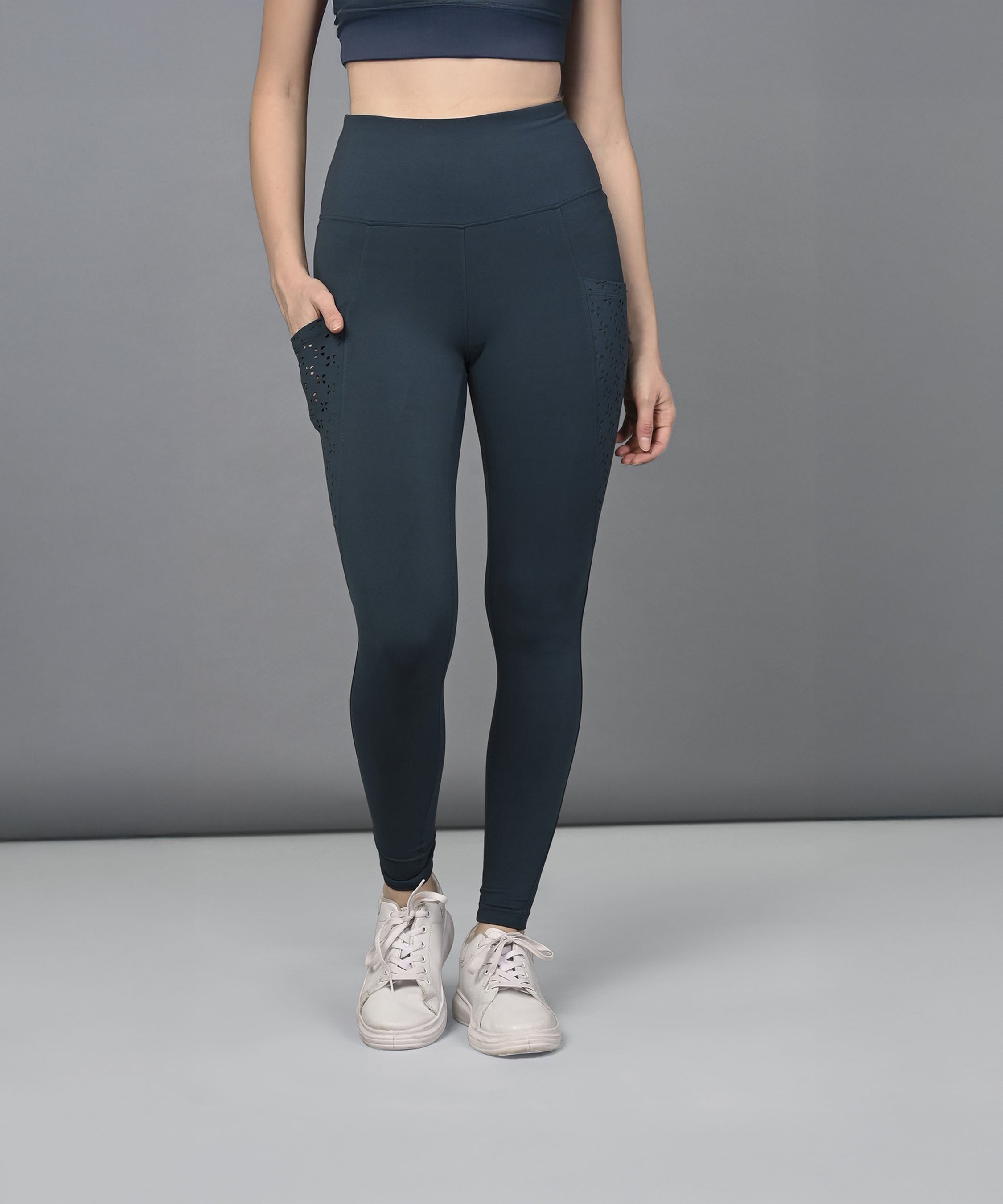 Woman in high-waisted teal leggings and white sneakers standing against a plain gray background.