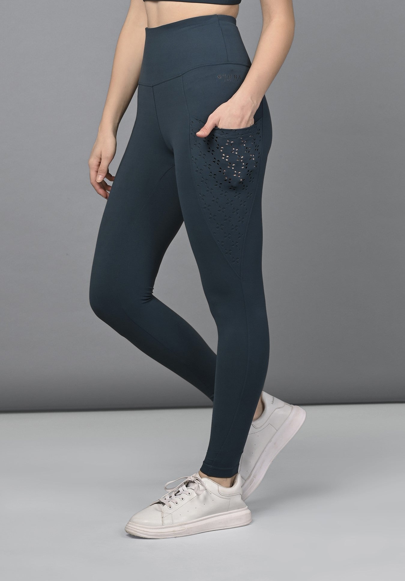 A woman wearing Teal Leggings featuring a high-waisted fit and laser-cut design, paired with white sneakers, stands against a gray background.