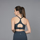 Woman wearing a teal sports bra and leggings stands with her back to the camera against a gray background.