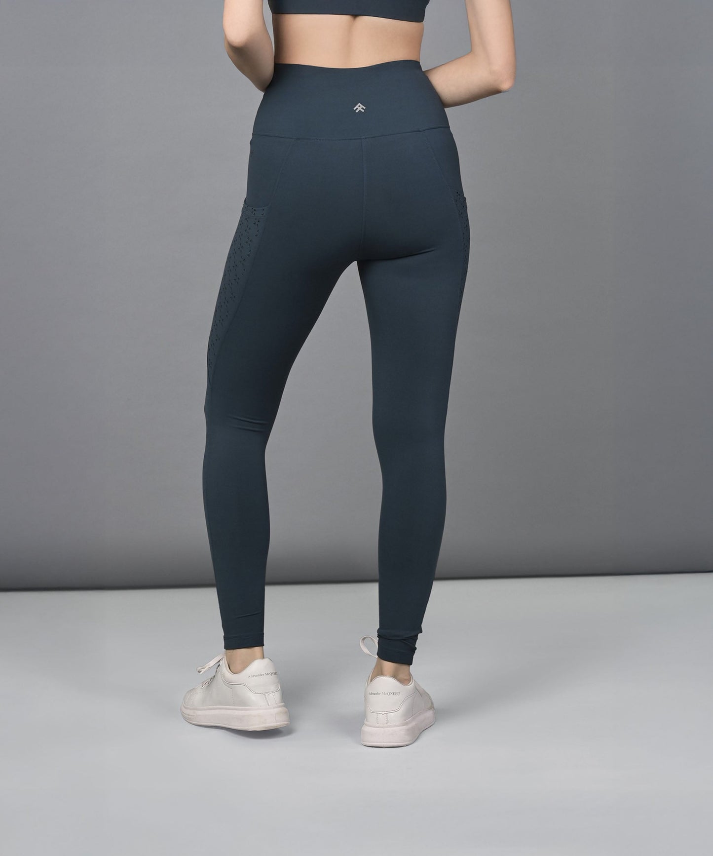 A woman wears teal leggings with a high-rise waistband and a sports bra, standing facing away in white sneakers against a gray background.
