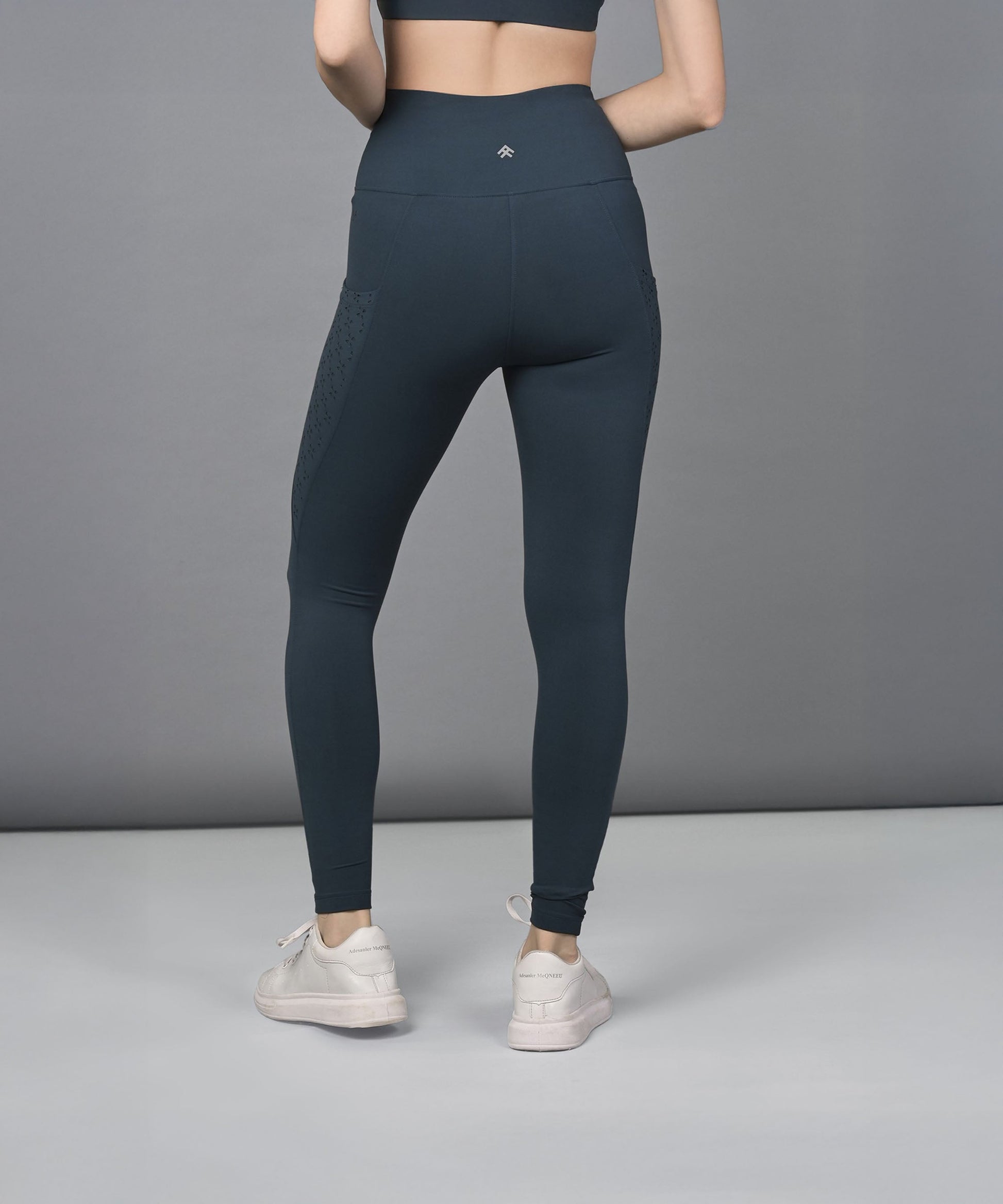 A woman wears teal leggings with a high-rise waistband and a sports bra, standing facing away in white sneakers against a gray background.