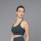 Woman in a teal sports bra and leggings stands against a plain gray background, looking to the side.