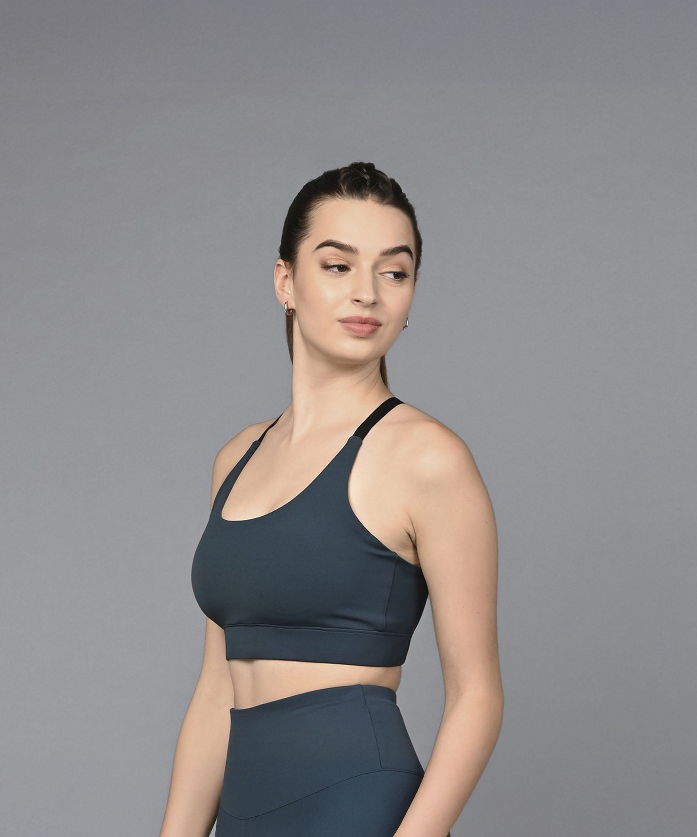 Woman in a teal sports bra and leggings stands against a plain gray background, looking to the side.