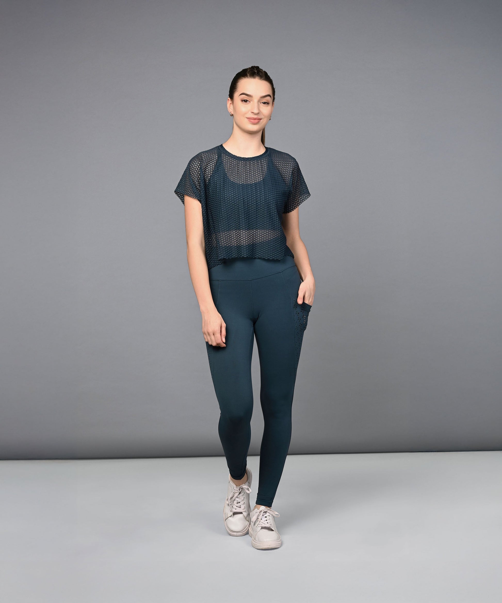 A woman stands against a plain gray background wearing teal leggings, a navy mesh top, and white sneakers.