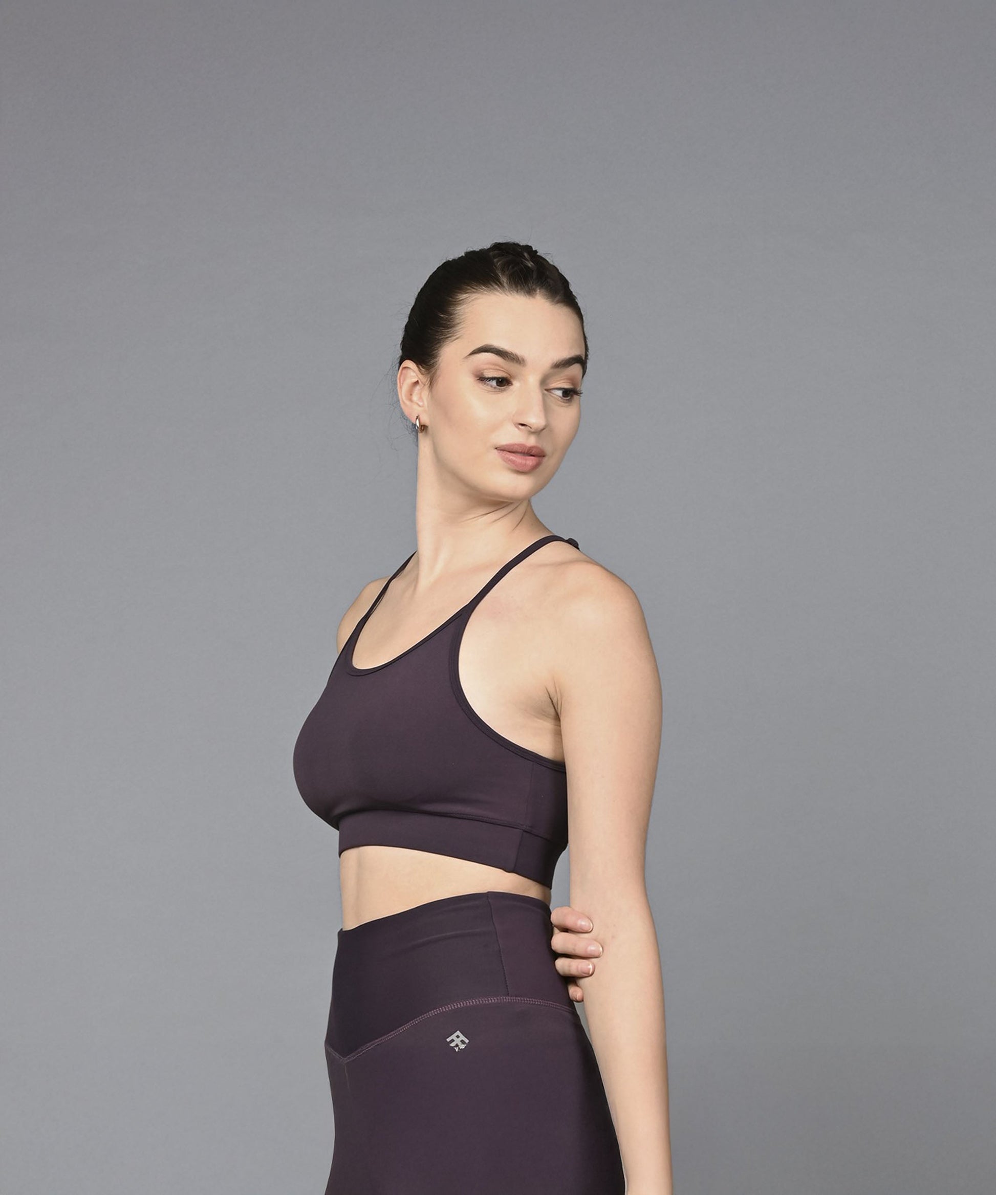 A woman in the Wine Sports Bra, featuring dark purple, moisture-wicking fabric, stands against a plain grey background and looks to the side.