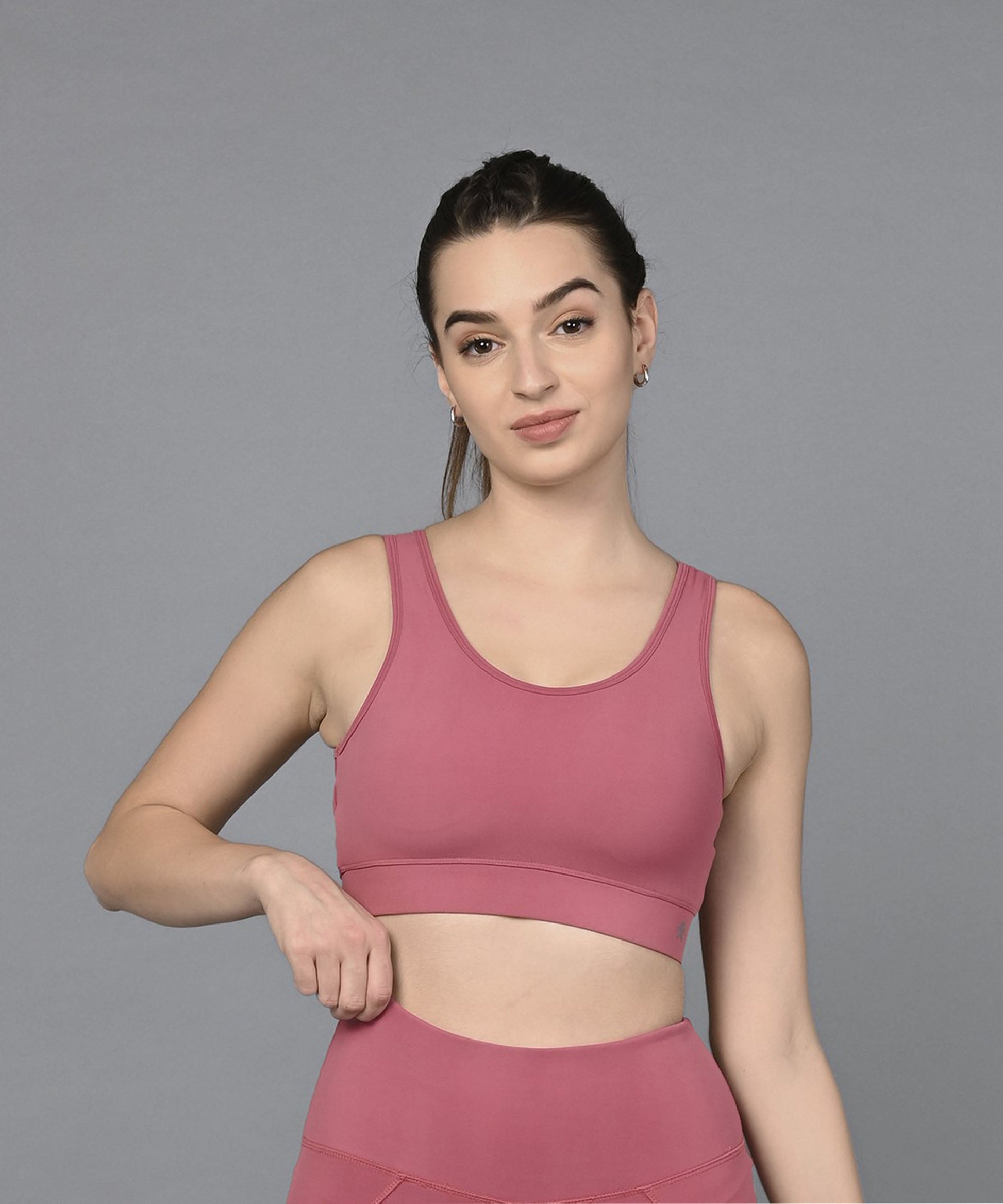 A woman in a Peony Pink Sports Bra and matching leggings stands against a gray background, holding the waistband of her moisture-wicking leggings.