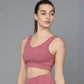 Wearing the Peony Pink Sports Bra, a woman stands against a gray background, looking to the side. The moisture-wicking design keeps her comfortable and stylish throughout any workout.