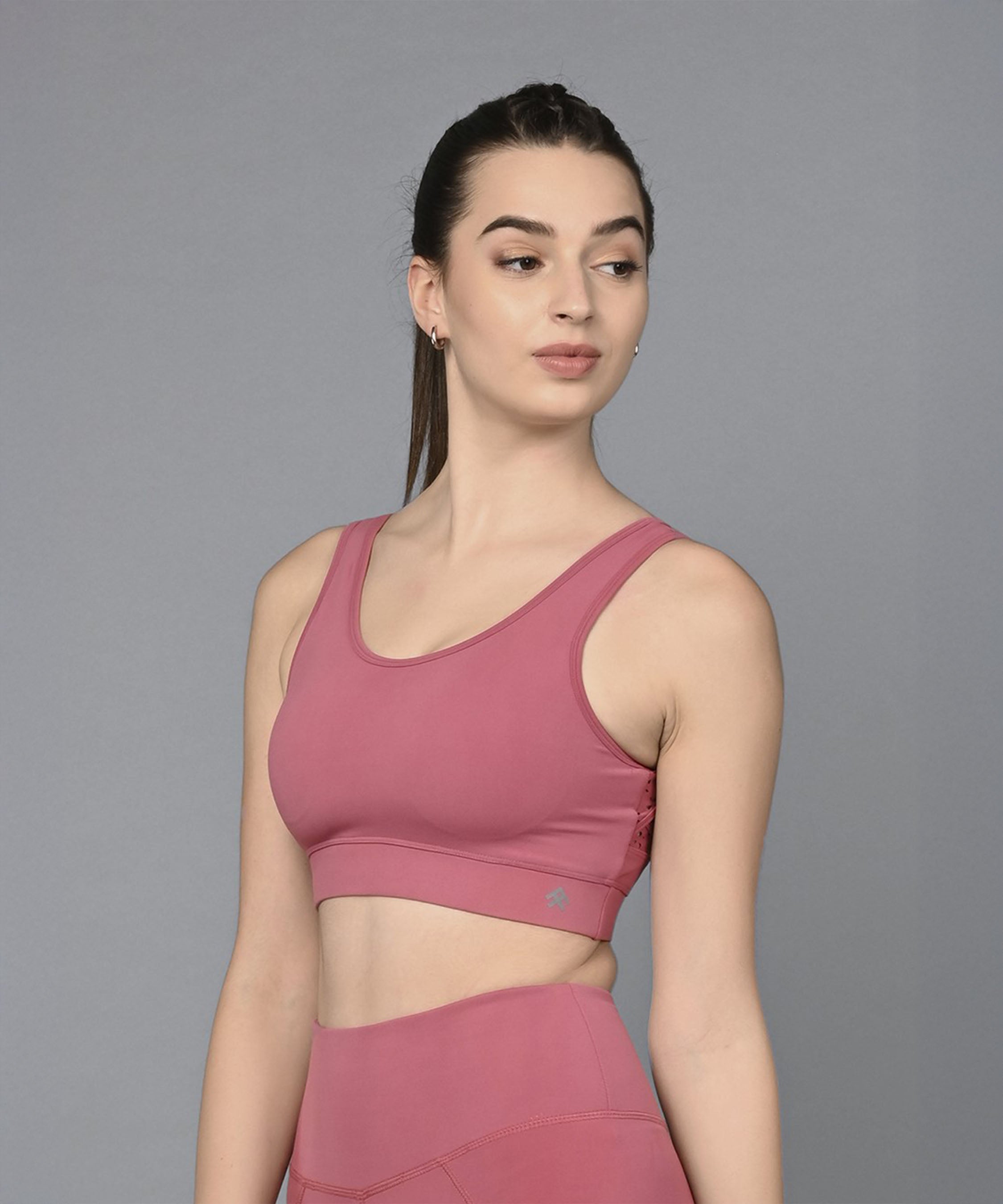 Wearing the Peony Pink Sports Bra, a woman stands against a gray background, looking to the side. The moisture-wicking design keeps her comfortable and stylish throughout any workout.