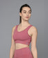 Wearing the Peony Pink Sports Bra, a woman stands against a gray background, looking to the side. The moisture-wicking design keeps her comfortable and stylish throughout any workout.
