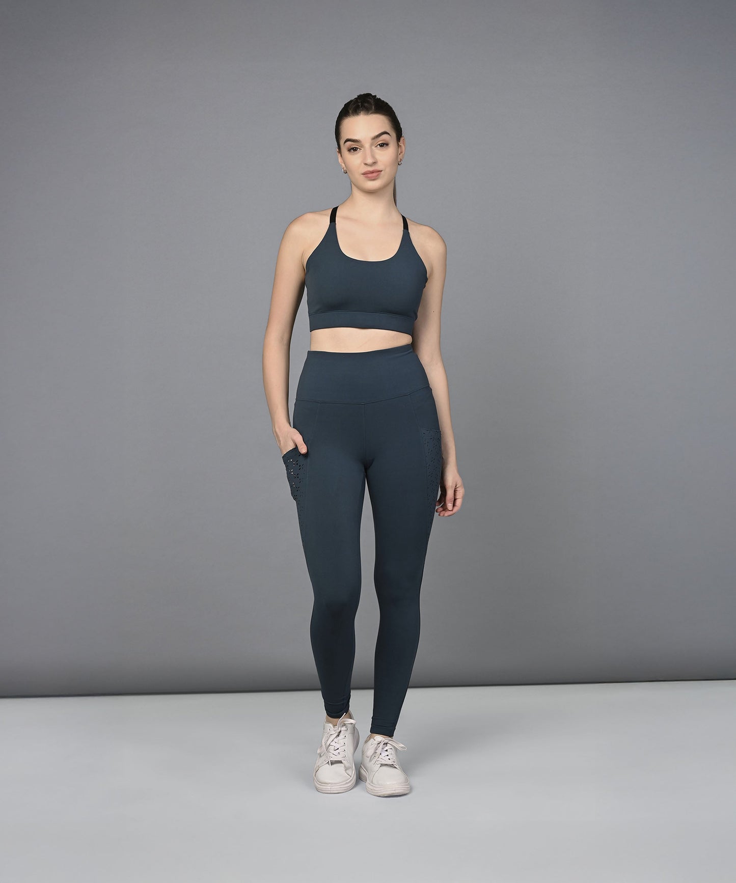 A woman models the Teal Netted Top with blue leggings, standing with one hand in her pocket against a gray background.