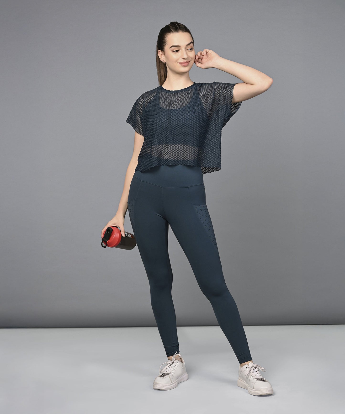 Woman wearing the Teal Netted Top, a stylish activewear piece with a honeycomb pattern, poses with a red water bottle against a plain gray background.