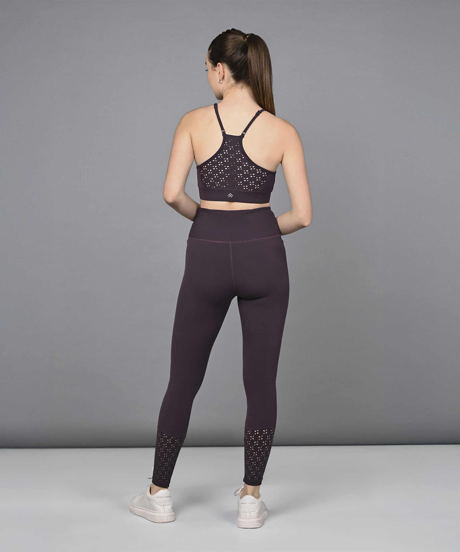 A woman stands with her back to the camera against a gray background, wearing the Wine Netted Top in dark purple athletic wear with a honeycomb self pattern and white sneakers.
