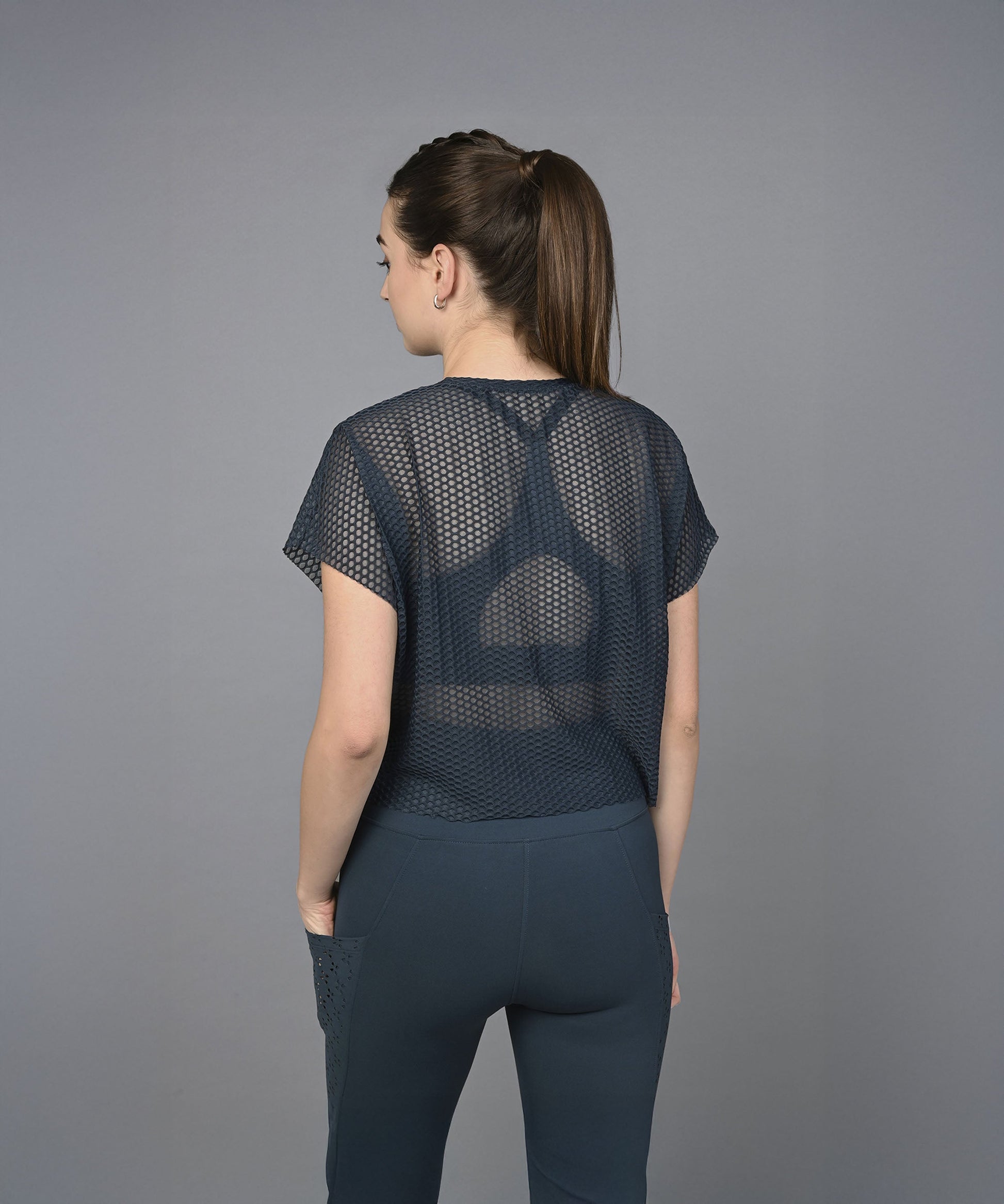 A woman with brown hair in a ponytail wears the Teal Netted Top, which features a subtle honeycomb self pattern, paired with matching pants and stands against a gray background.