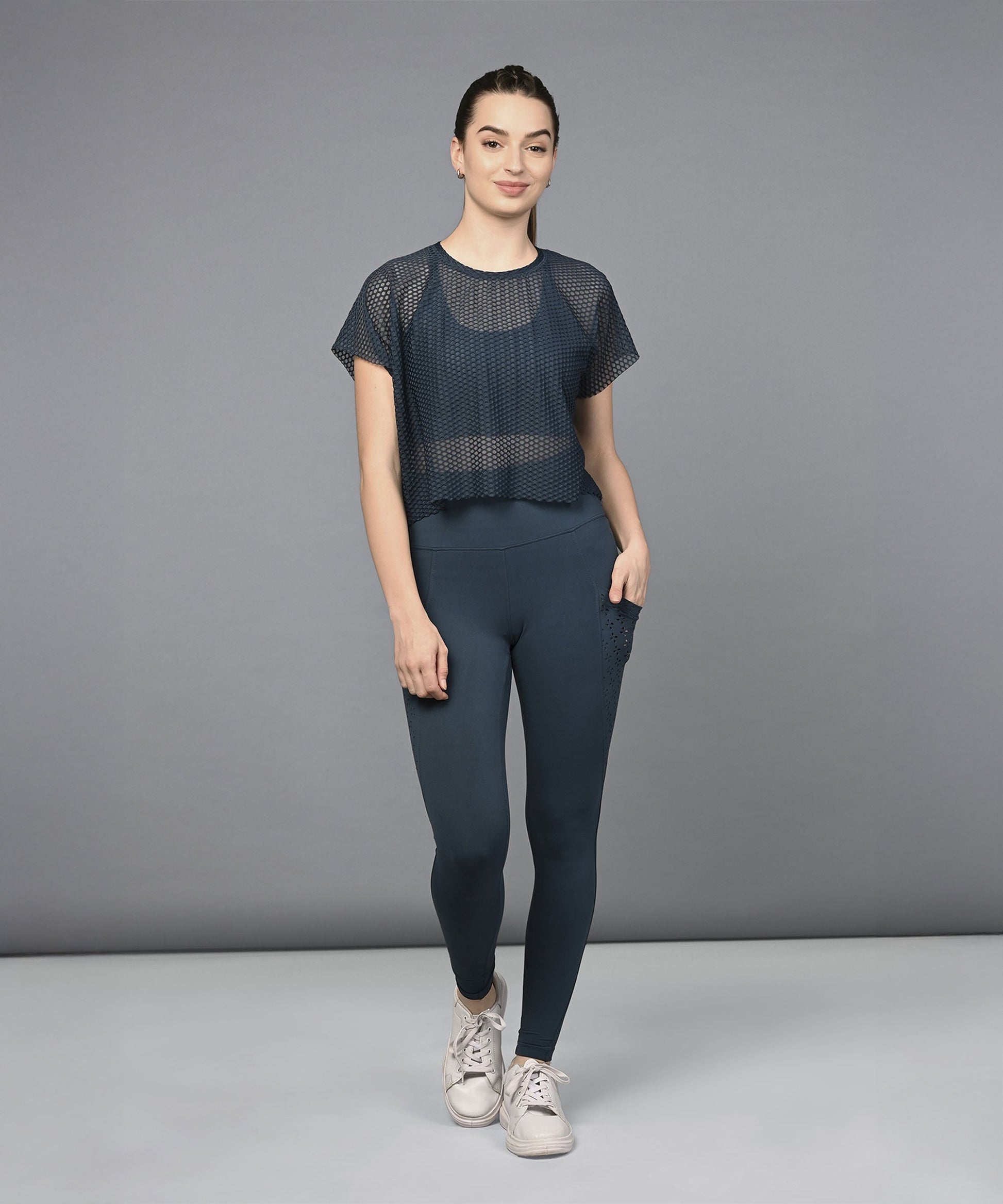 Woman wearing a Teal Netted Top with high-waisted leggings and sneakers, standing against a plain gray background.
