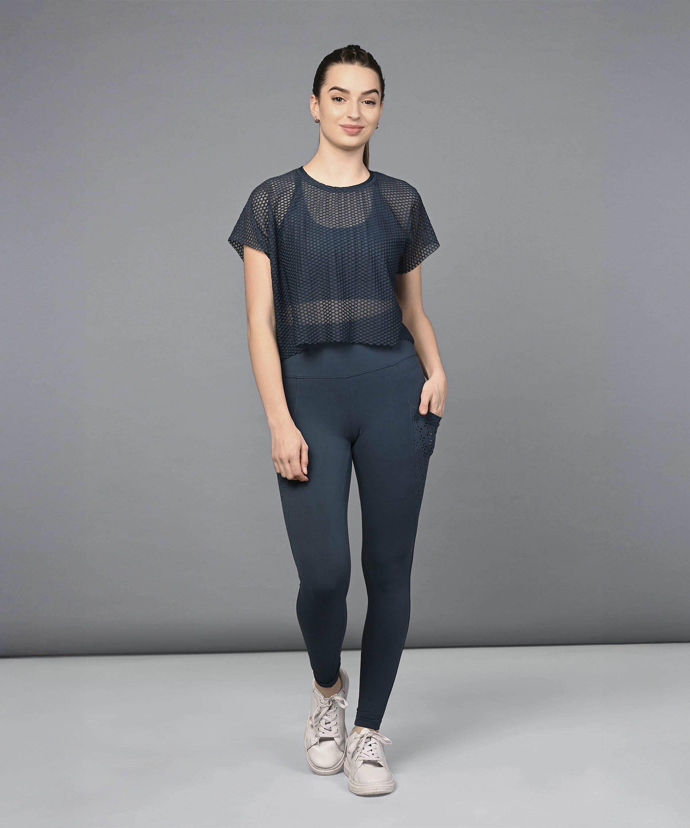 Woman wearing a Teal Netted Top with high-waisted leggings and sneakers, standing against a plain gray background.