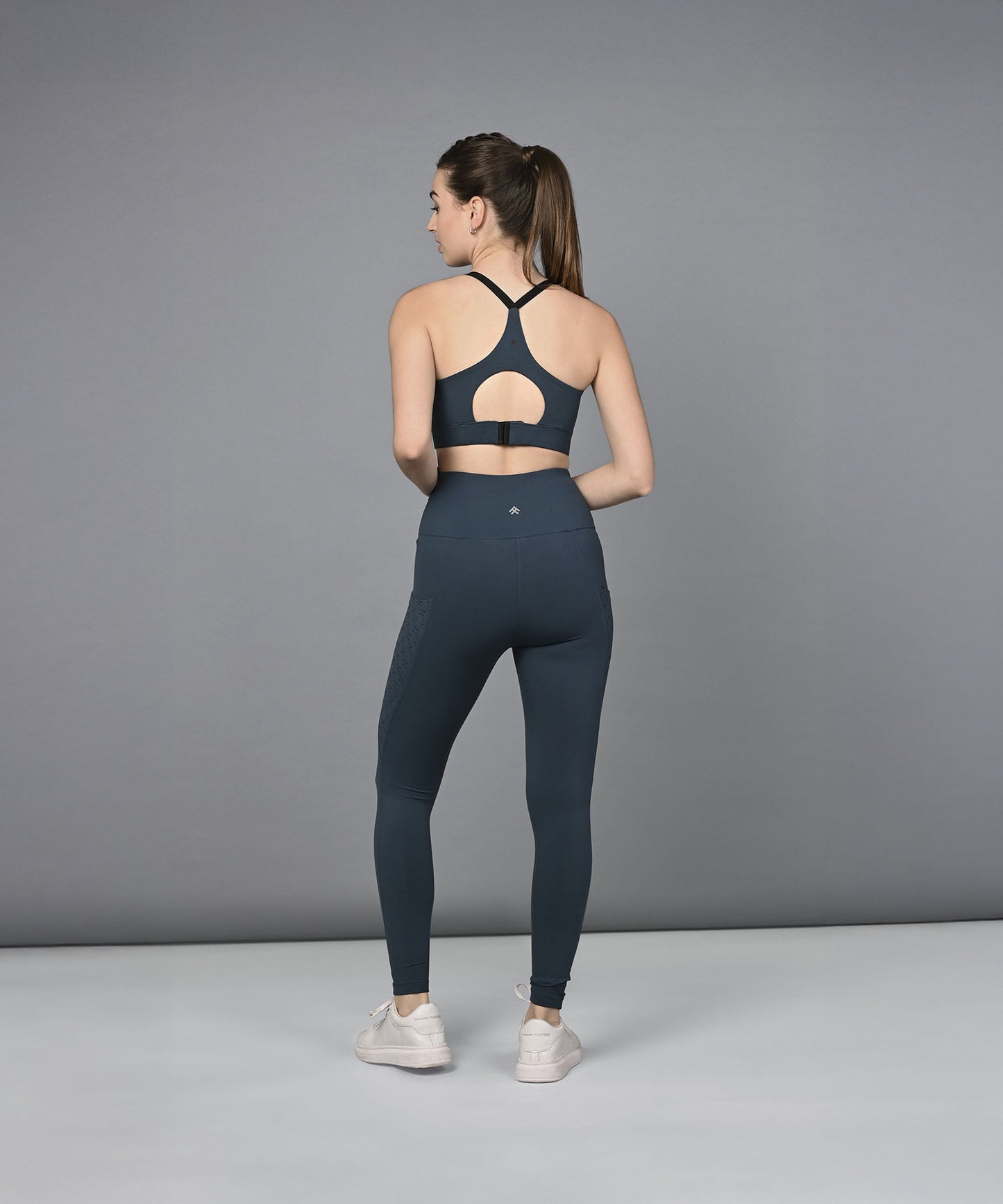 Woman in a Teal Netted Top and leggings stands facing away on a gray background.