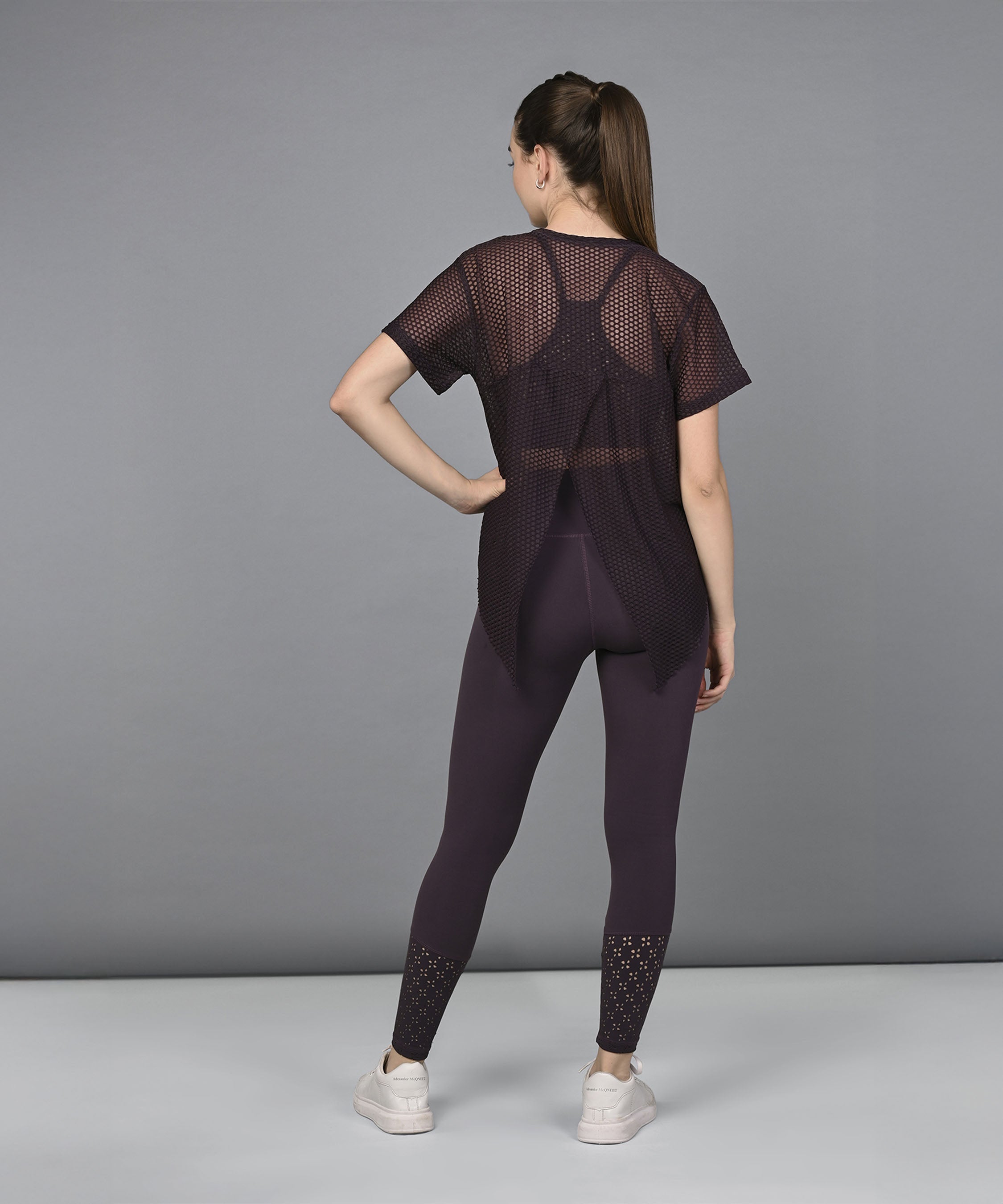 A woman wearing the Wine Netted Top with a honeycomb pattern, paired with leggings and sneakers, stands facing away against a plain gray background.