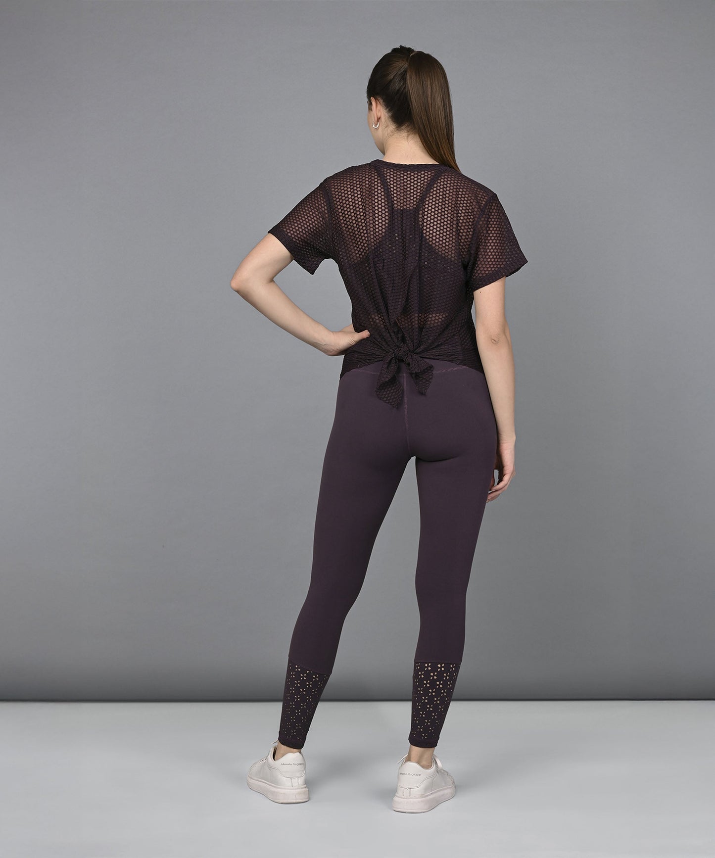 Woman in activewear stands facing away, wearing a sexy Wine Netted Top, purple leggings, and white sneakers.