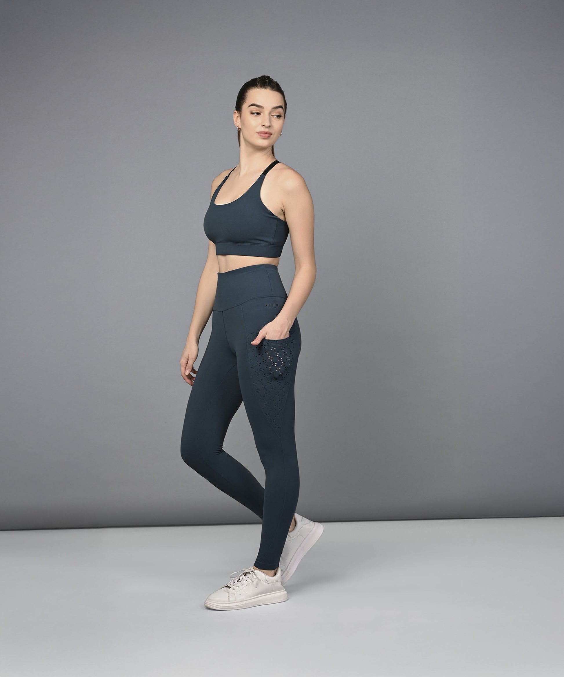 A woman poses against a gray background wearing the Teal Netted Top, honeycomb pattern leggings, and white sneakers.