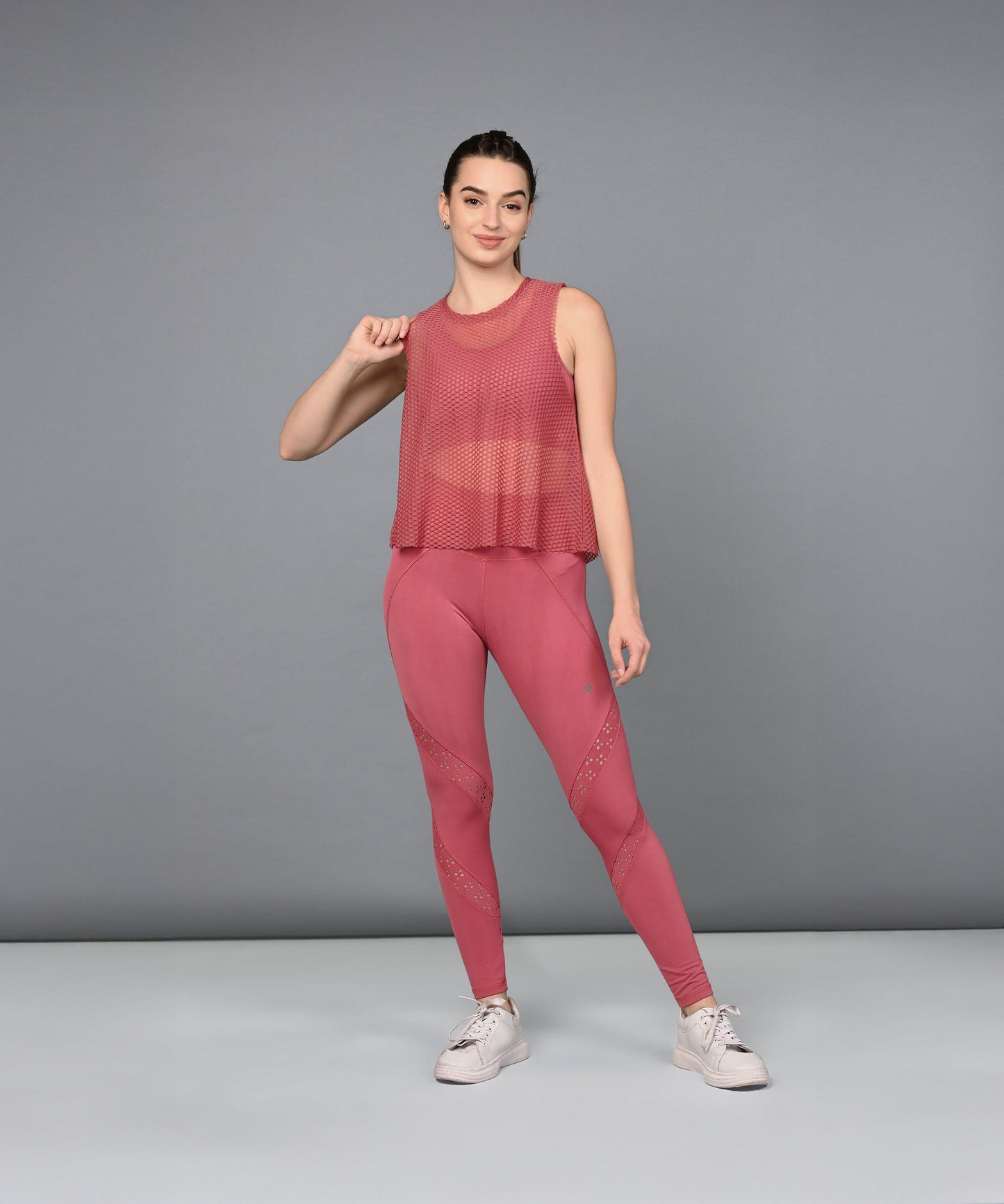 A woman models Peony Pink Leggings with a pink mesh tank top, posing gracefully against a plain gray background.