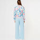 A woman faces away on a plain white background, wearing the Blossom Blue Set with Crop Top featuring light blue cargo track pants and a floral bomber jacket.
