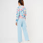 A woman faces away wearing the Blossom Blue Set with Crop Top—floral bomber jacket, light blue cargo track pants, and white shoes—against a white background.