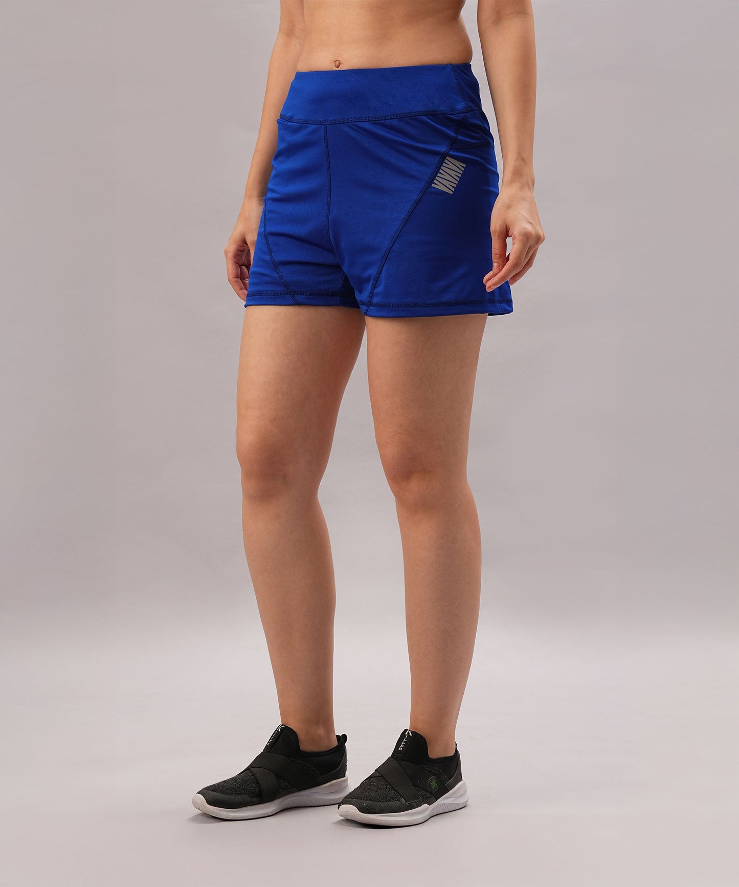 A woman stands against a plain light background wearing Blue Premium Stretch Shorts With Lining, paired with black slip-on sneakers.