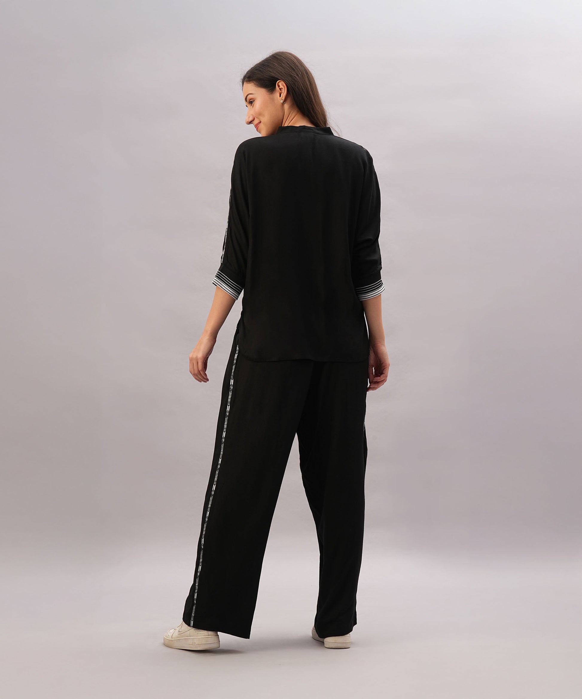 A woman faces backward against a plain light background, wearing the Black Rayon Co-Ord Set featuring a stylish top and loose-fitting pants, paired with white sneakers.
