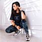 A woman in casual wear squats indoors by a sunlit white wall, wearing the Positive Body Image T Shirt, a black basic tee that celebrates confidence and self-love.