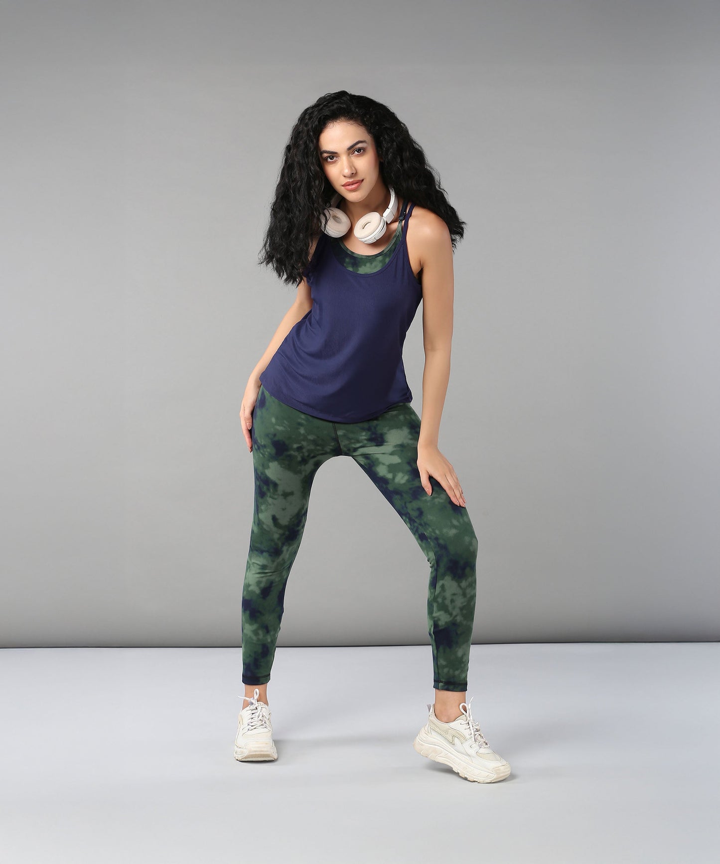 A woman with curly hair wears headphones, a Tie & Dye Bra T shirt, green leggings, and white sneakers as she poses confidently.