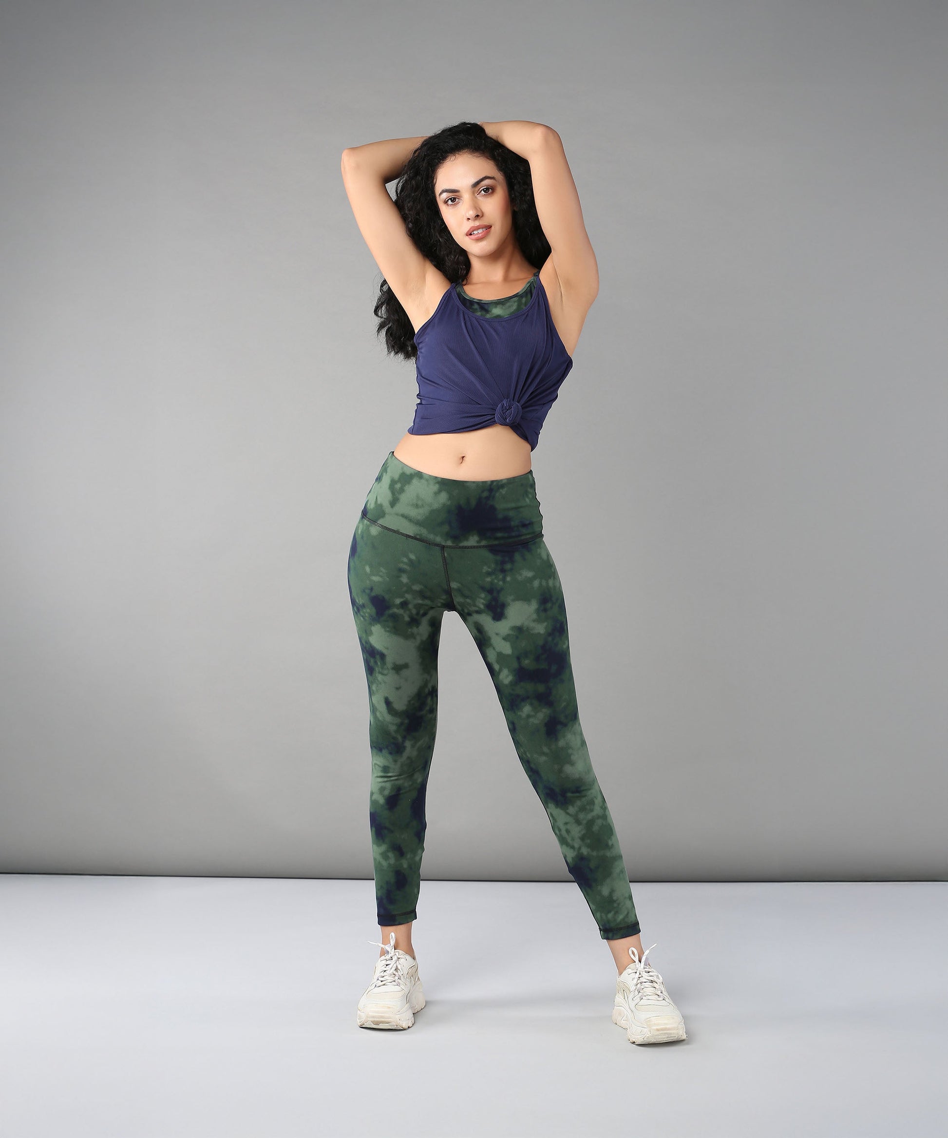 Woman poses with arms raised, wearing the Tie & Dye Bra T shirt in blue—featuring a built-in bra—paired with green tie-dye leggings and white sneakers.