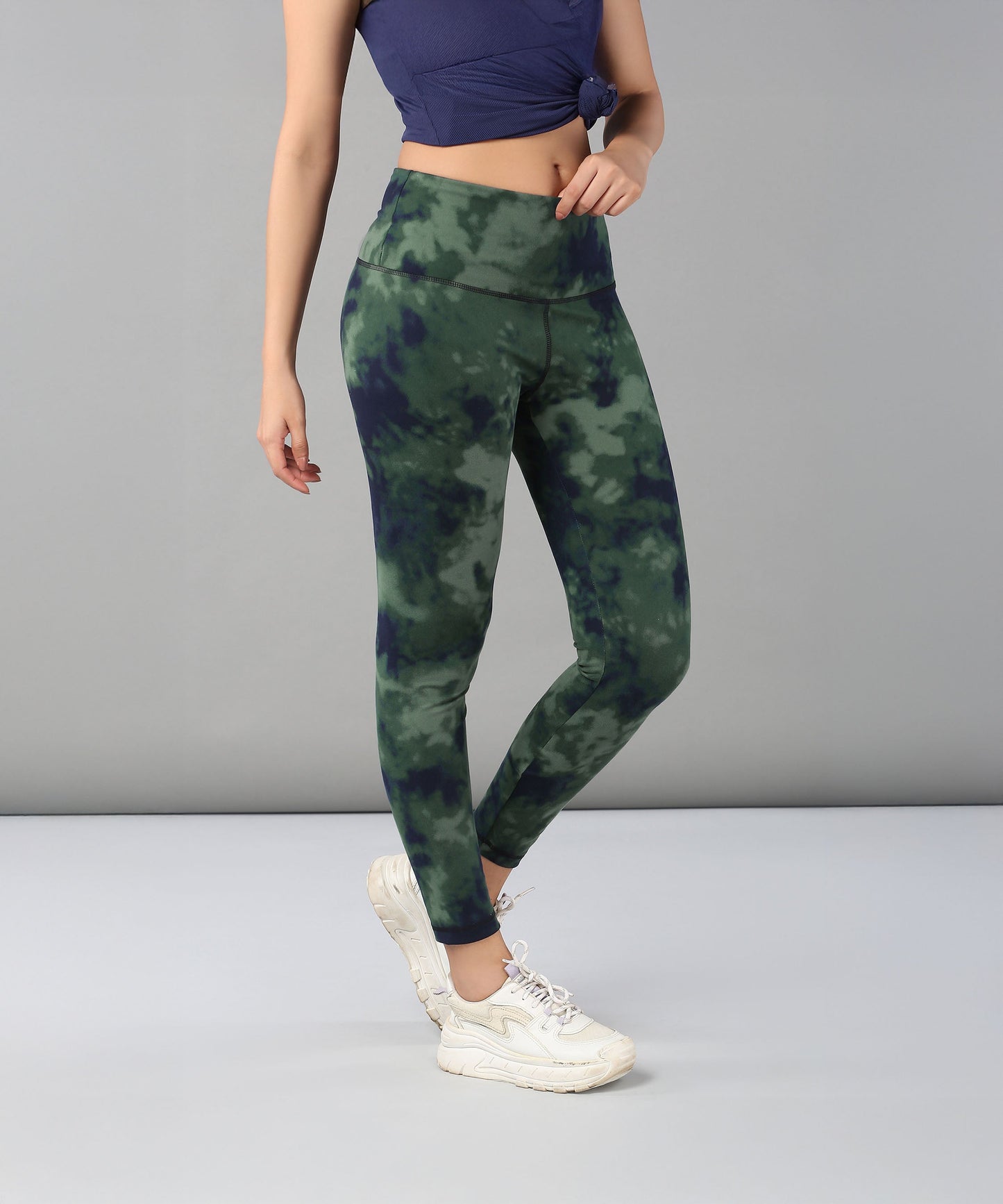 A woman models Tie & Dye Leggings in green stretchy fabric, paired with white sneakers and a blue top, standing against a plain gray background—ideal for enhancing your workout wardrobe.